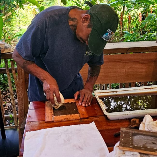 A man is using a cloth to wipe a wooden frame filled with a brown substance on a table surrounded by greenery.