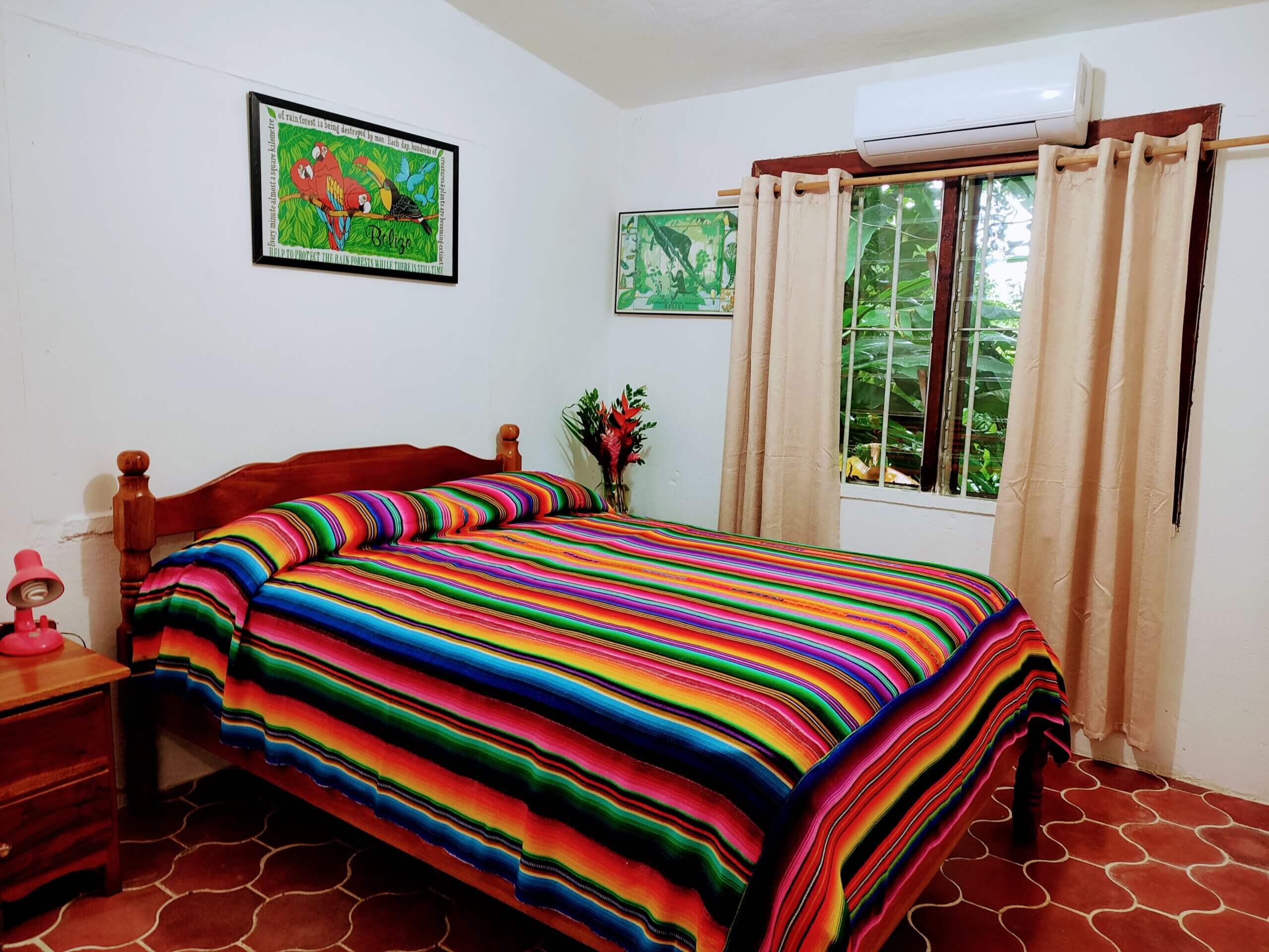 A bedroom with white walls and red patterned tile flooring, featuring a bed covered in a vibrant multi-colored striped blanket and a window with neutral tan curtains.