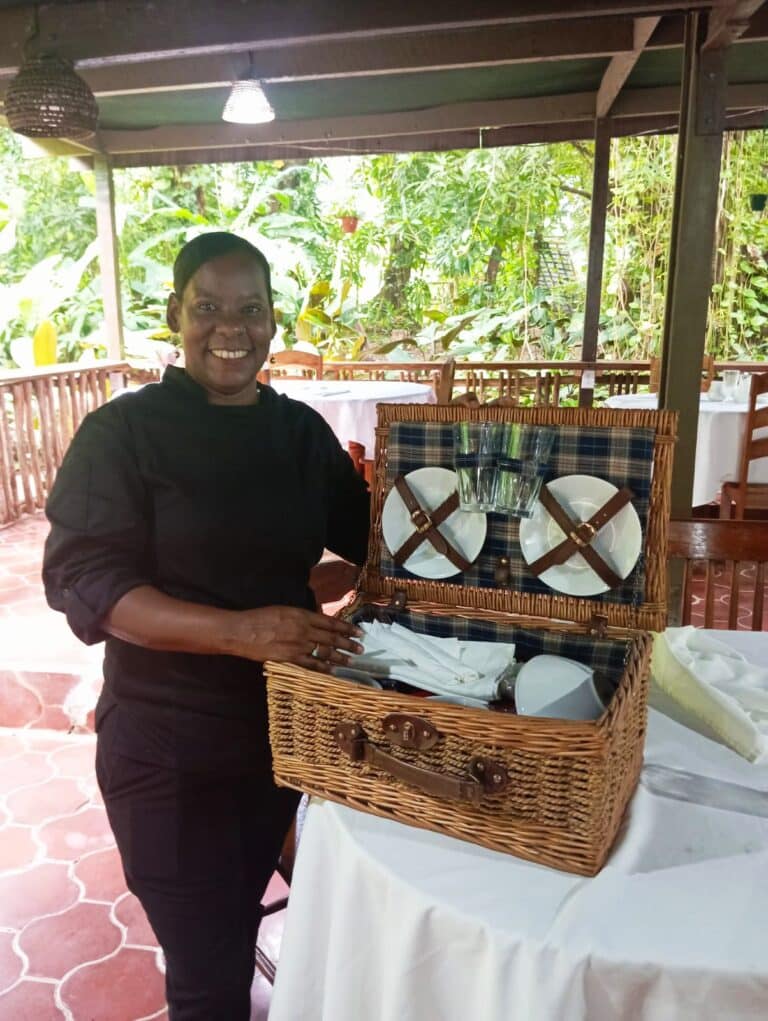 A smiling woman in a black outfit stands beside an open picnic basket filled with dishes and utensils in a lush restaurant setting.