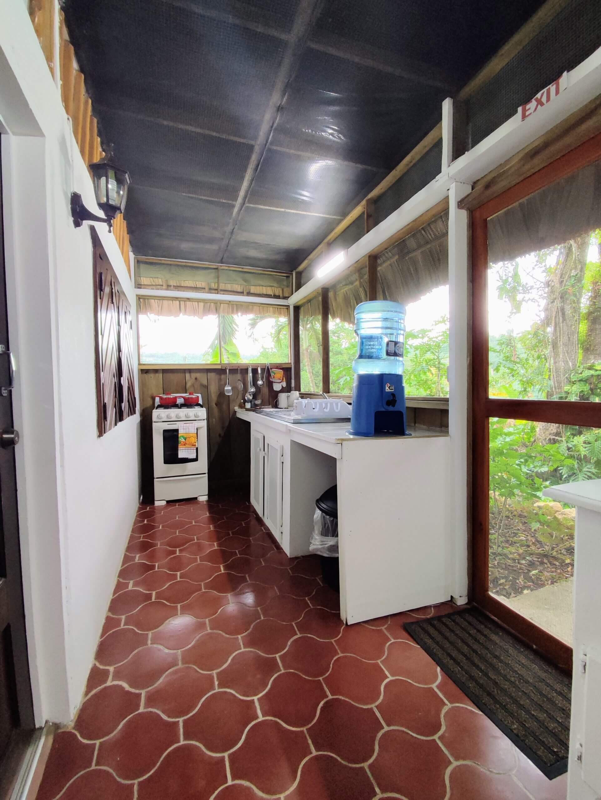 A narrow outdoor-style kitchen or porch with red tile floors, white counters, a water cooler, and a screened door leading to a wooded area.