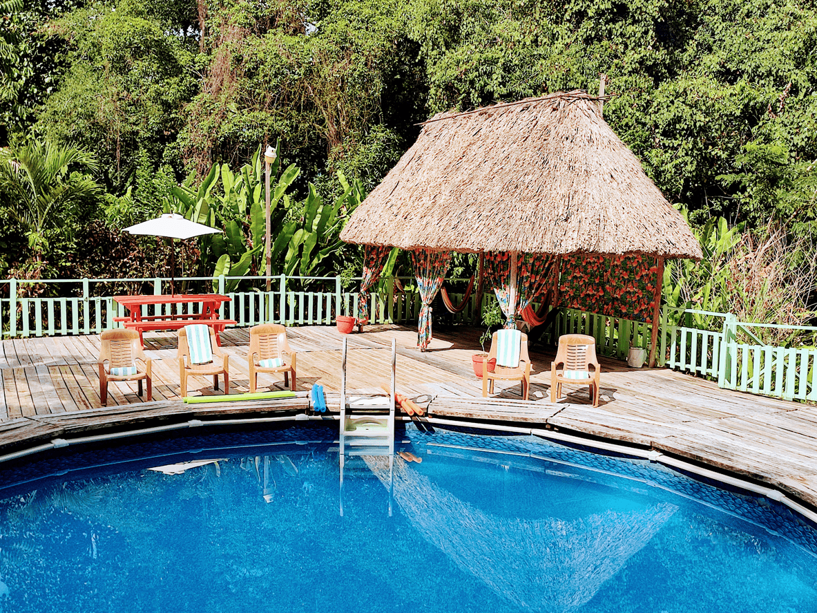 A tranquil poolside scene featuring a thatched hut, sun loungers, and lush greenery in the background.