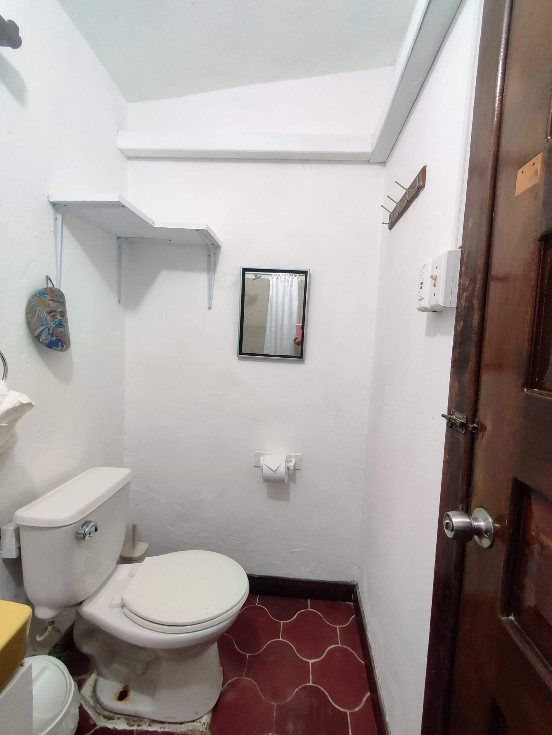 A simple bathroom with white walls and red patterned tile flooring, featuring a white toilet and a small framed mirror hanging on the wall.