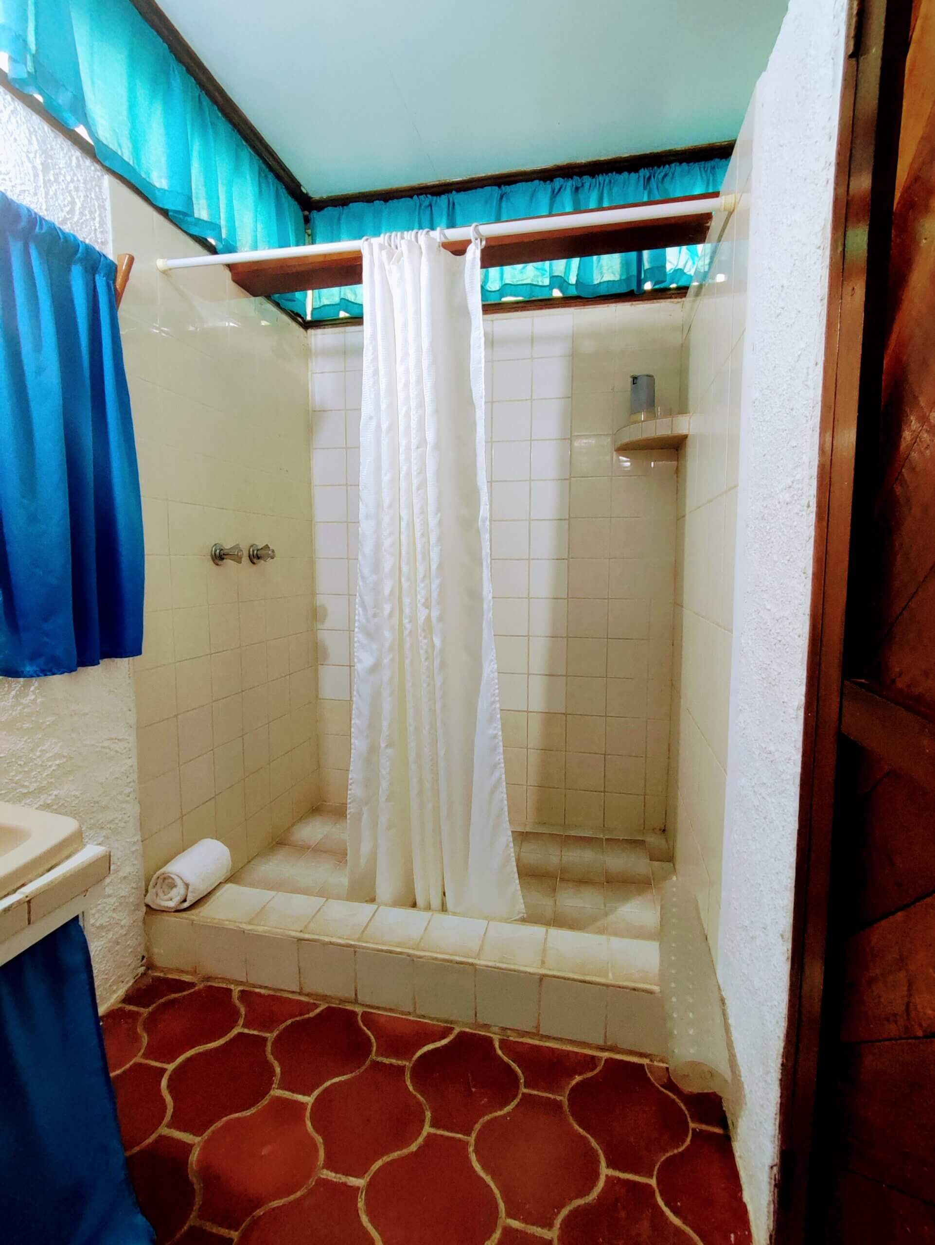 A walk-in shower with white tiled walls and a white shower curtain, featuring red patterned tile flooring and bright blue curtains visible in the upper corner.