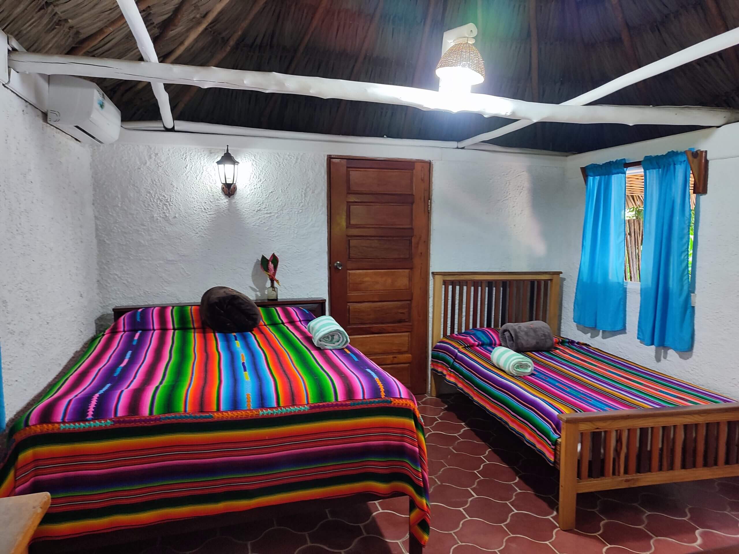 A bedroom with white walls and red patterned tile flooring, featuring two beds covered in vibrant multi-colored striped blankets, a wooden door, and a window with bright blue curtains.