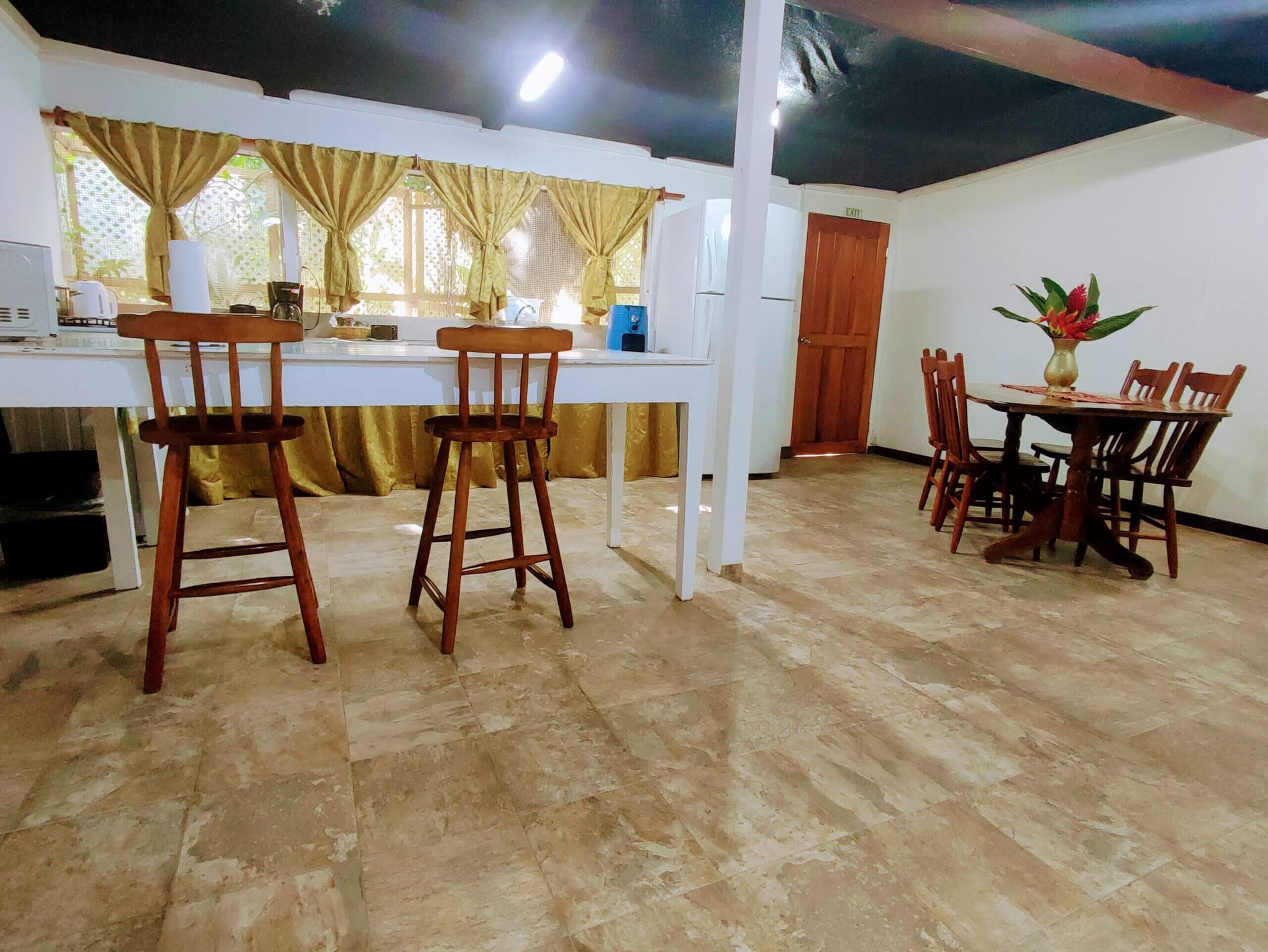 A spacious dining and kitchen area with light-colored tile flooring, featuring a wooden table with chairs, a white island counter with two wooden barstools, and windows with gold-colored curtains.