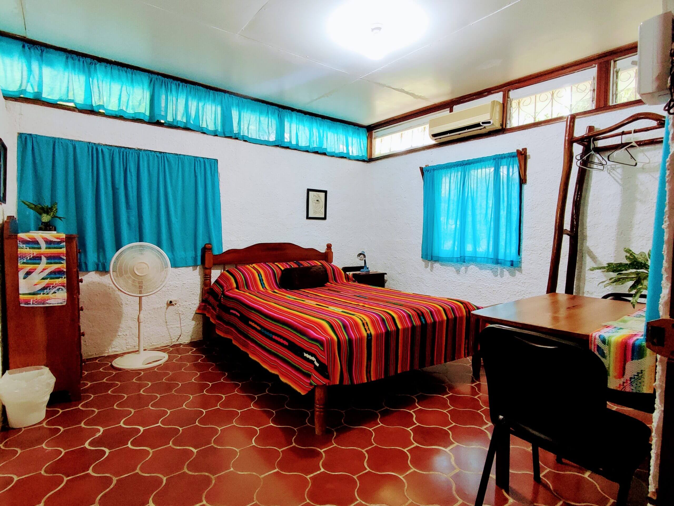 A bedroom with white walls and red patterned tile flooring, featuring a bed with a vibrant multi-colored striped blanket, a white pedestal fan, and windows with bright teal curtains.