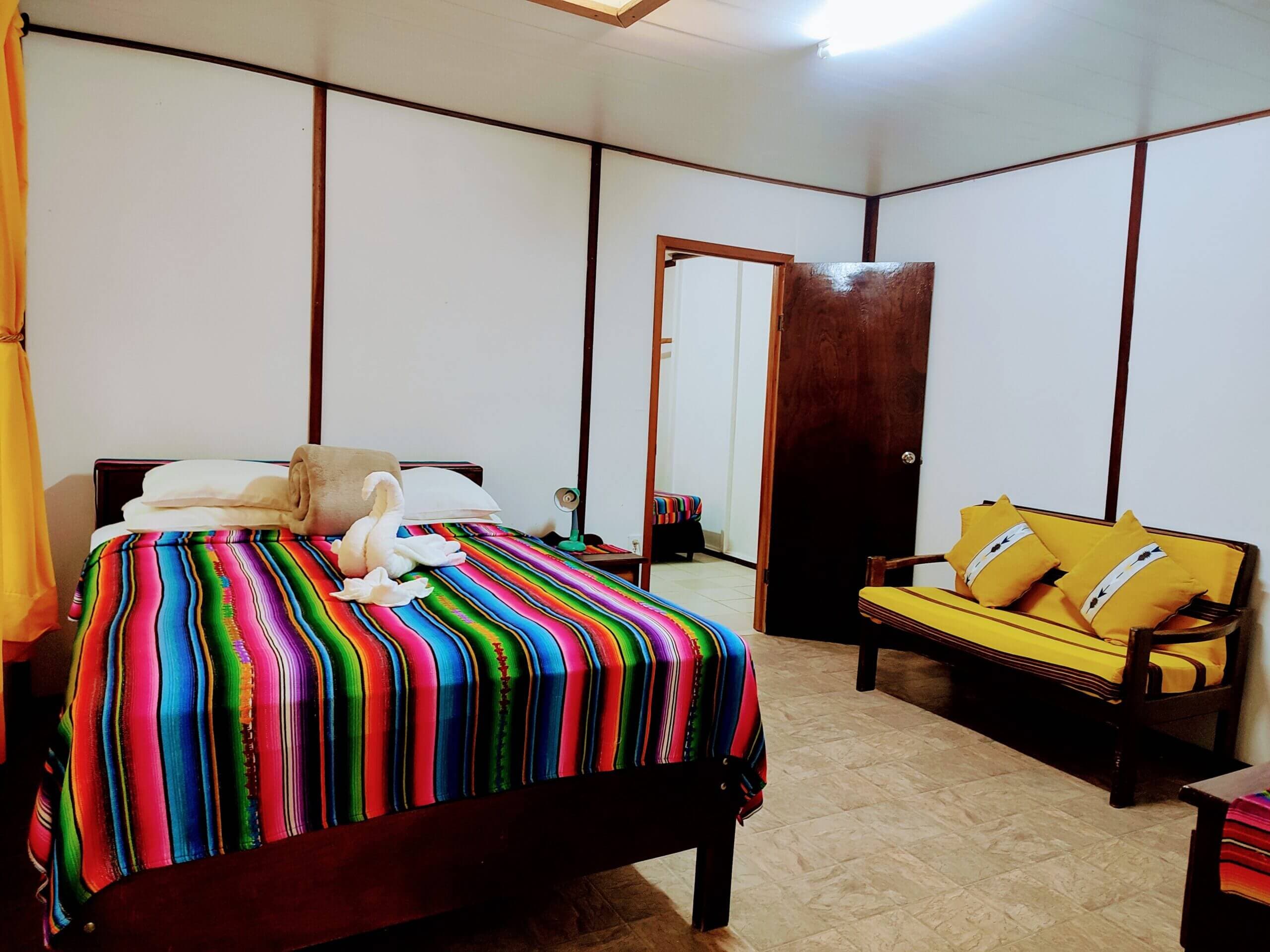 A bedroom with white paneled walls and light-colored flooring, featuring a bed with a vibrant multi-colored striped blanket and a wooden sofa with yellow cushions and patterned throw pillows.