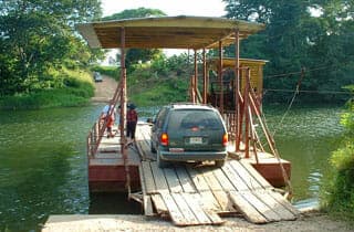 A vehicle drives onto a wooden ferry crossing a river surrounded by greenery.
