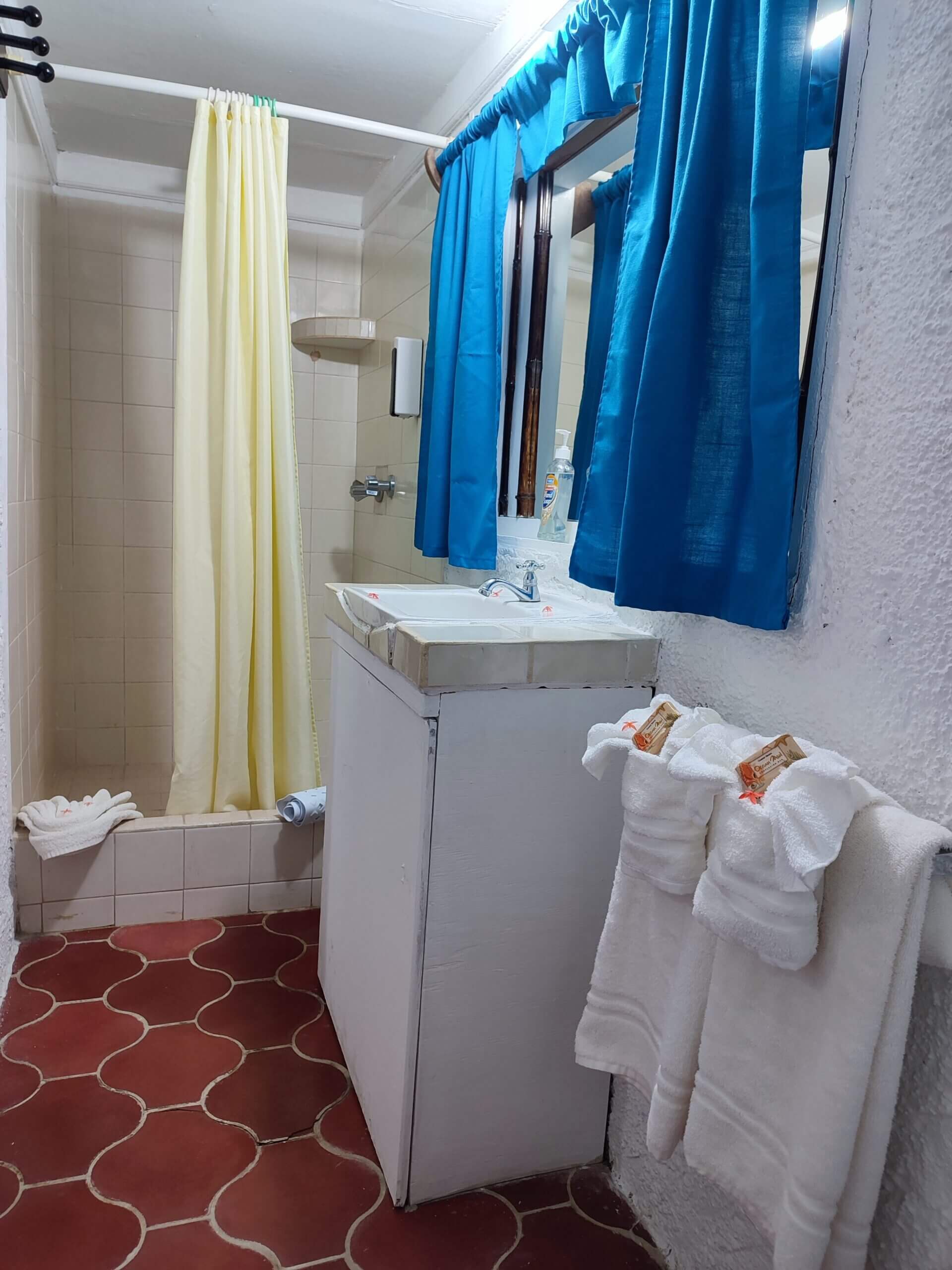 A bathroom with red patterned tile flooring, featuring a white pedestal sink, bright blue curtains over a mirror, and a shower area with a pale yellow curtain.
