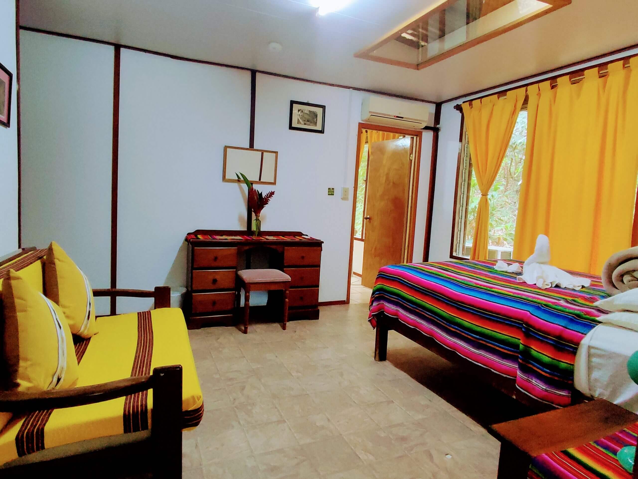 A bedroom with white paneled walls and light-colored tile flooring, featuring a bed with a vibrant multi-colored striped blanket, a wooden desk with a small chair, and a wooden sofa with yellow cushions and matching yellow curtains on the window.