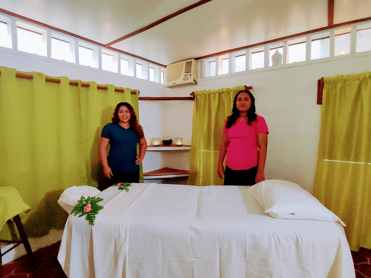 Two women stand in a massage therapy room beside a prepared massage table, with green curtains in the background.