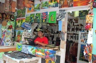 A man sits at a counter in a vibrant art shop filled with colorful paintings and crafts.