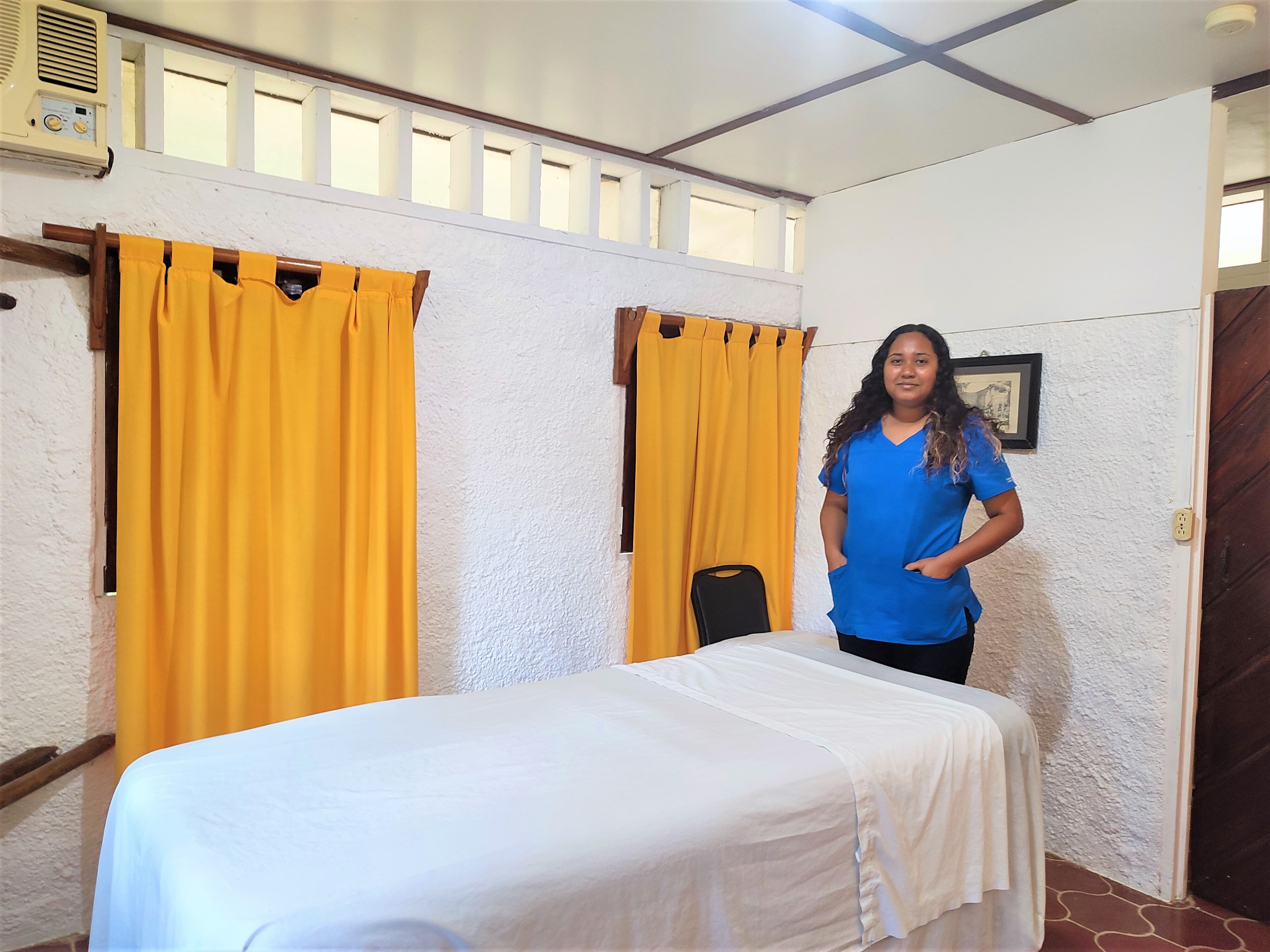 A woman in a blue shirt stands beside a massage table in a softly lit room with yellow curtains.