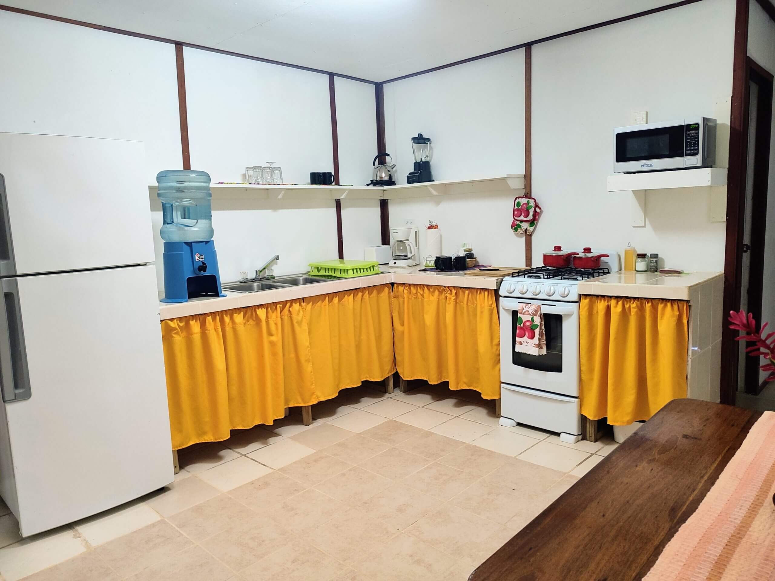 A spacious kitchen with white walls and light-colored tile flooring, featuring L-shaped white countertops with yellow fabric skirts, a white refrigerator, a white gas stove, and a blue water cooler.