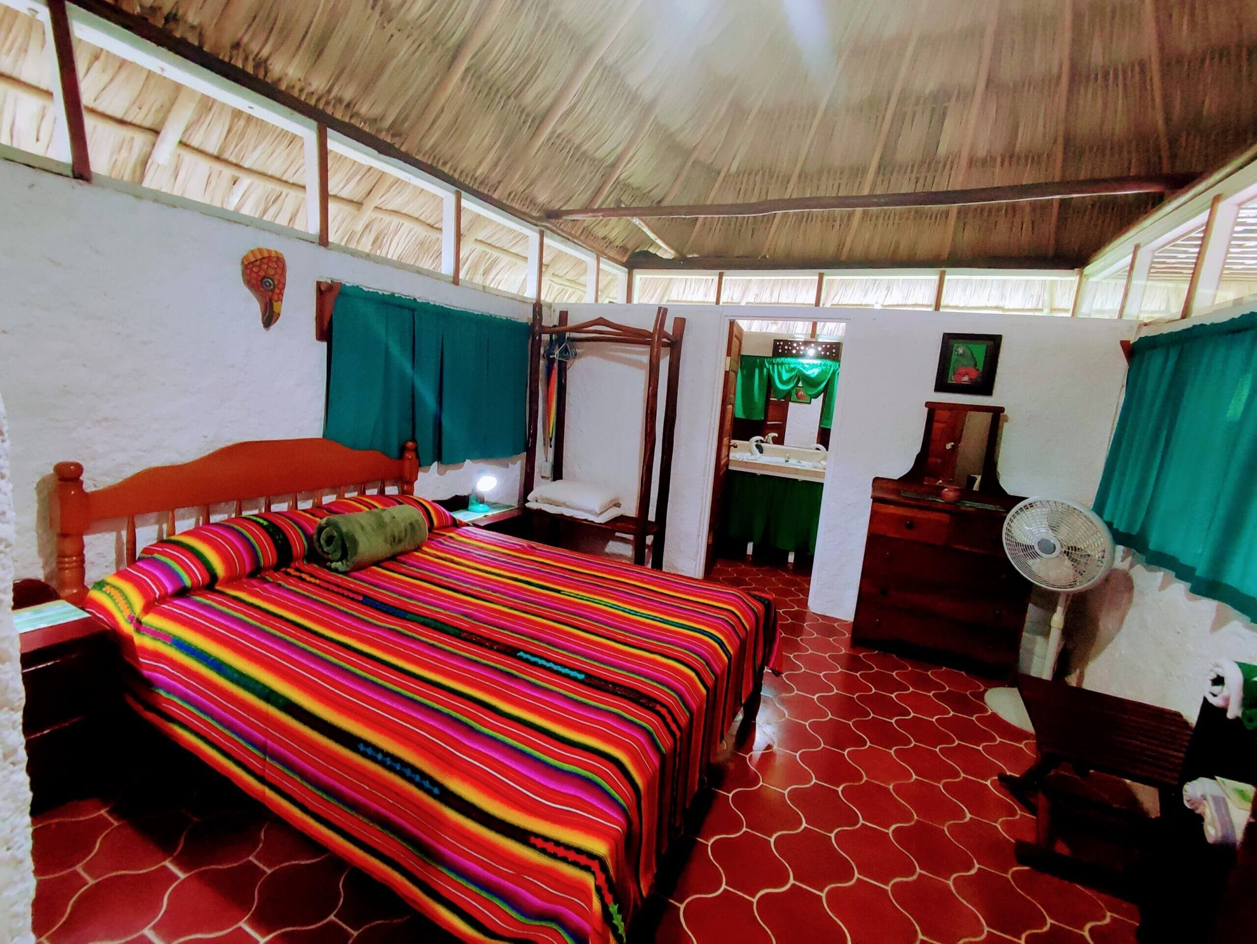 A bedroom with red patterned tile flooring and a vaulted wooden ceiling, featuring a large bed with a vibrant multi-colored striped blanket, dark wood furniture, and windows with deep green curtains.
