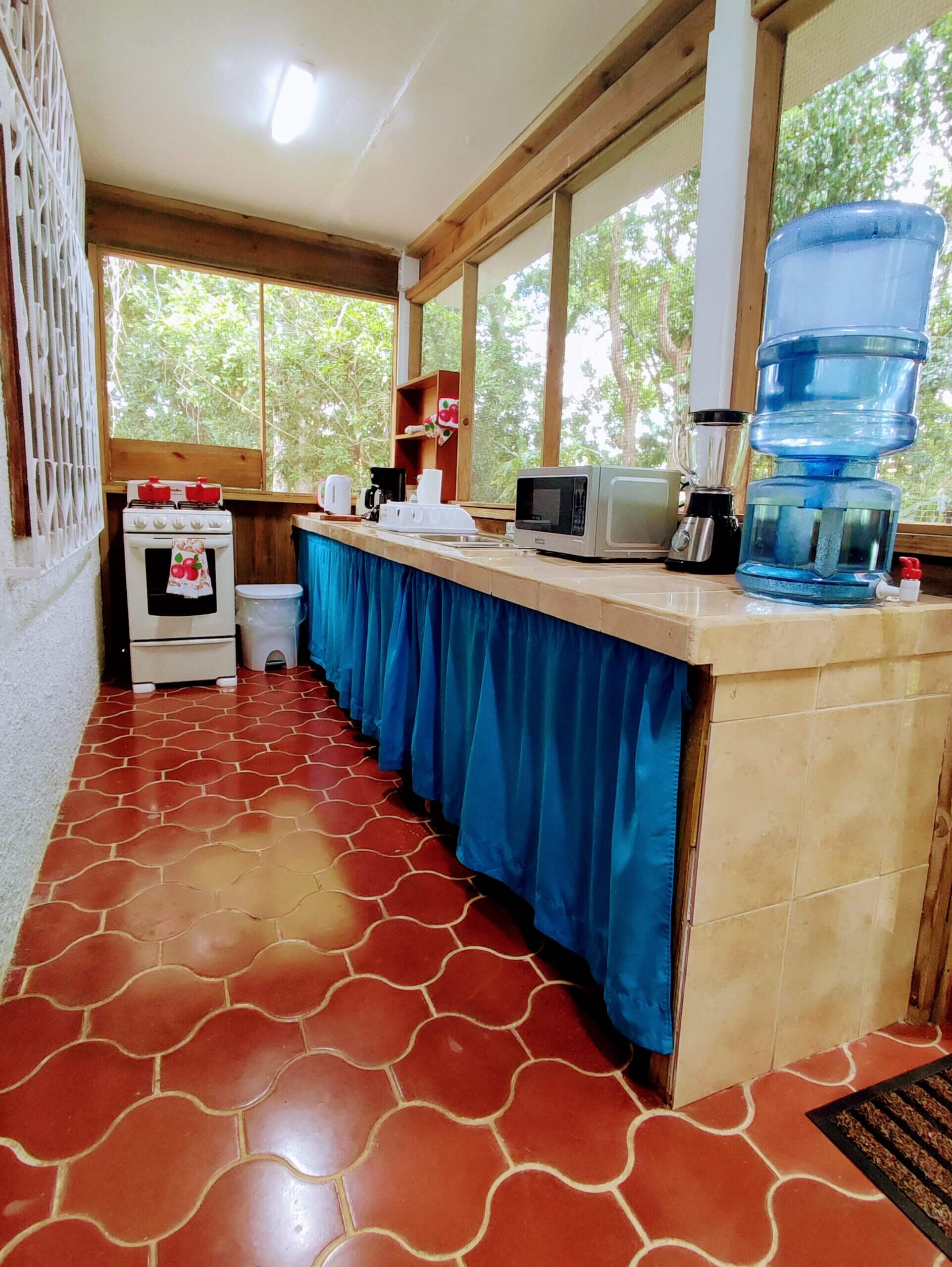 A long, narrow indoor kitchenette with red patterned tile flooring, featuring a white stove at the far end and a long tan counter with a bright blue fabric skirt, equipped with a microwave and a water cooler.