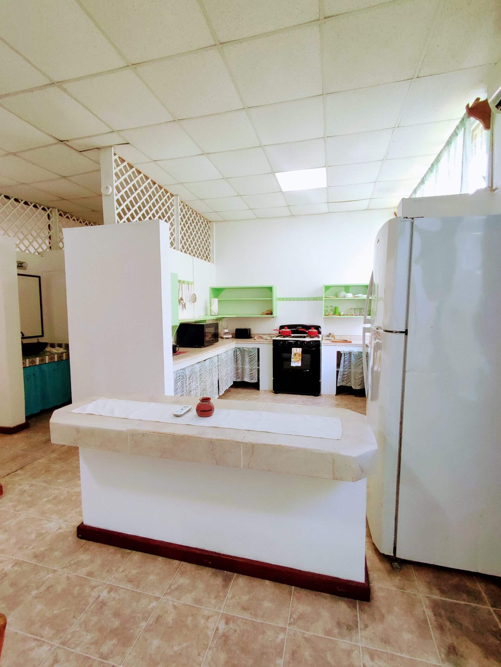 An open-concept kitchen area with white walls and light-colored tile flooring, featuring a white island counter, a full-sized white refrigerator, and a black stove in the background.