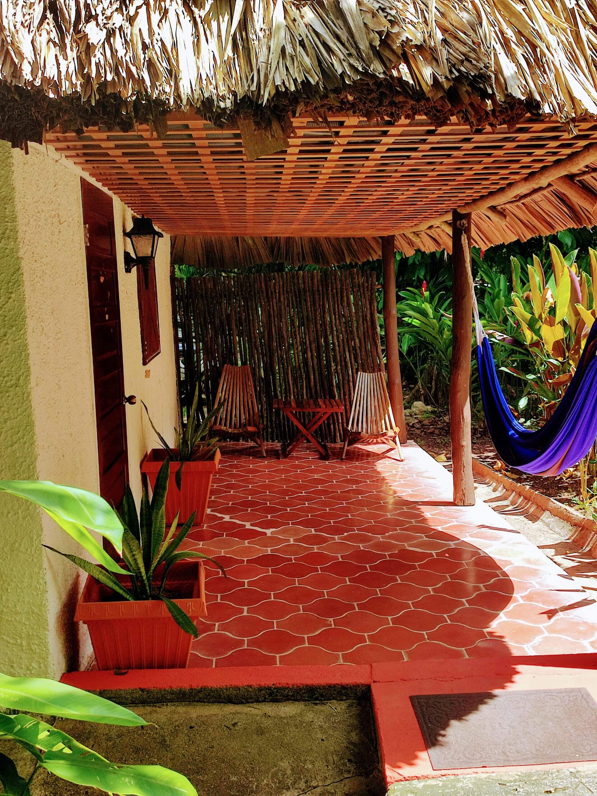 A red patterned tile patio with a thatched roof, featuring two wooden lounge chairs, a blue hammock, and potted plants.