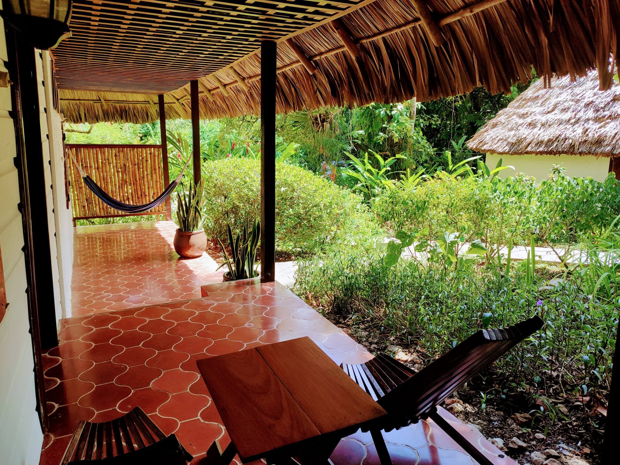 An outdoor covered patio with red patterned tile flooring, featuring wooden chairs, a small table, and a brown hammock, overlooking a lush green garden under a thatched roof.