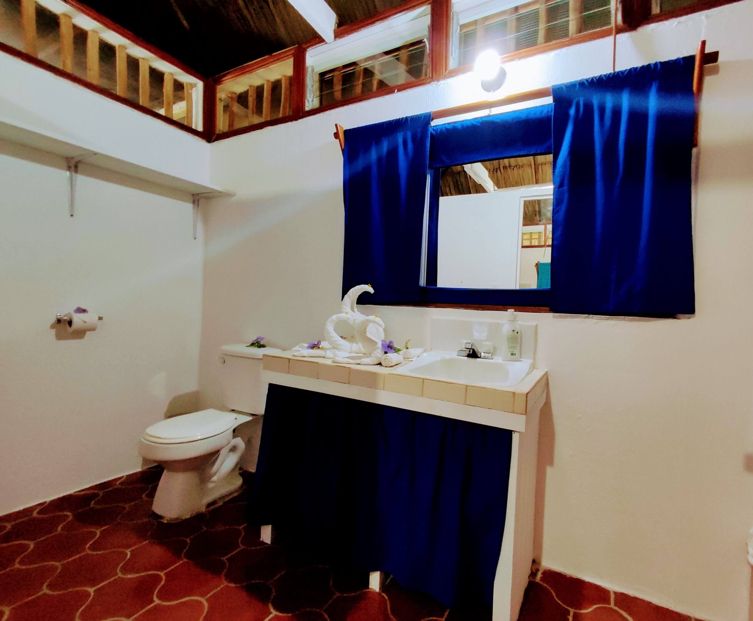 A bathroom featuring a white vanity with a dark blue fabric skirt, a white toilet, and dark blue curtains over a window, all set against white walls and red patterned tile flooring.