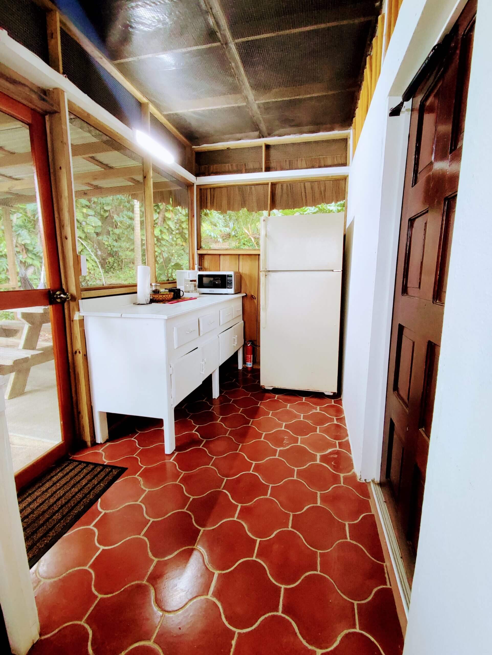 A kitchenette with red patterned tile flooring, a white refrigerator, a white kitchen island with a microwave, and a wooden door on the right.