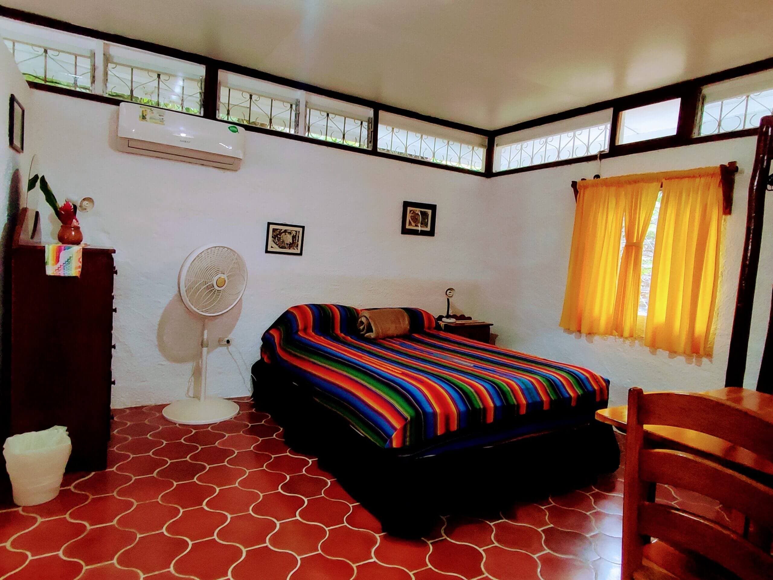 A bedroom with white walls and red patterned tile flooring, featuring a bed with a vibrant multi-colored striped blanket, a white pedestal fan, and a window with yellow curtains.
