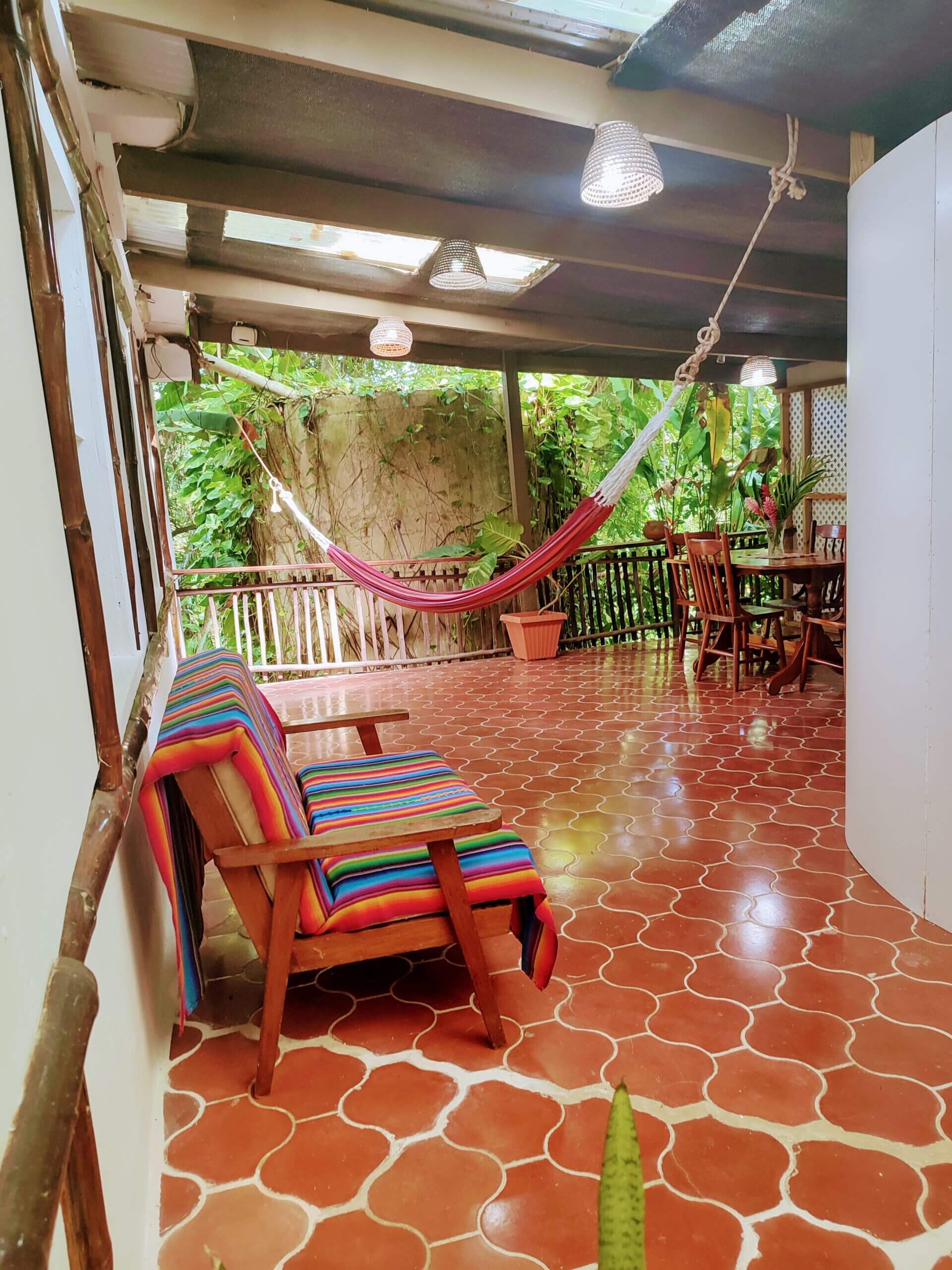 An outdoor patio with red patterned tile flooring, featuring a wooden chair with a vibrant multi-colored striped cushion and a red hammock strung across the space, overlooking a lush green garden.