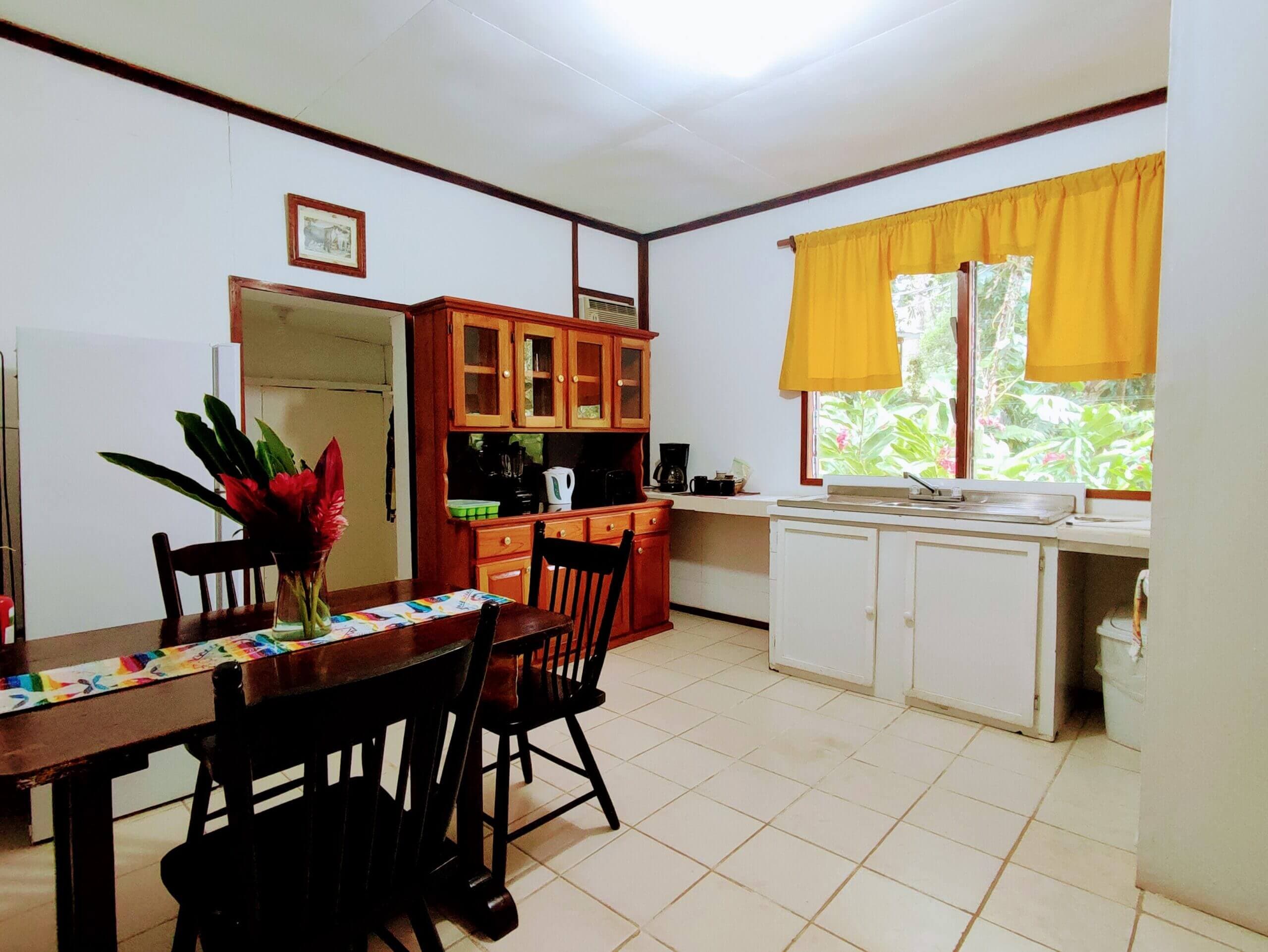 A spacious kitchen and dining area featuring a wooden dining table with black chairs, a large wooden hutch, white cabinetry, and a window with bright yellow curtains.