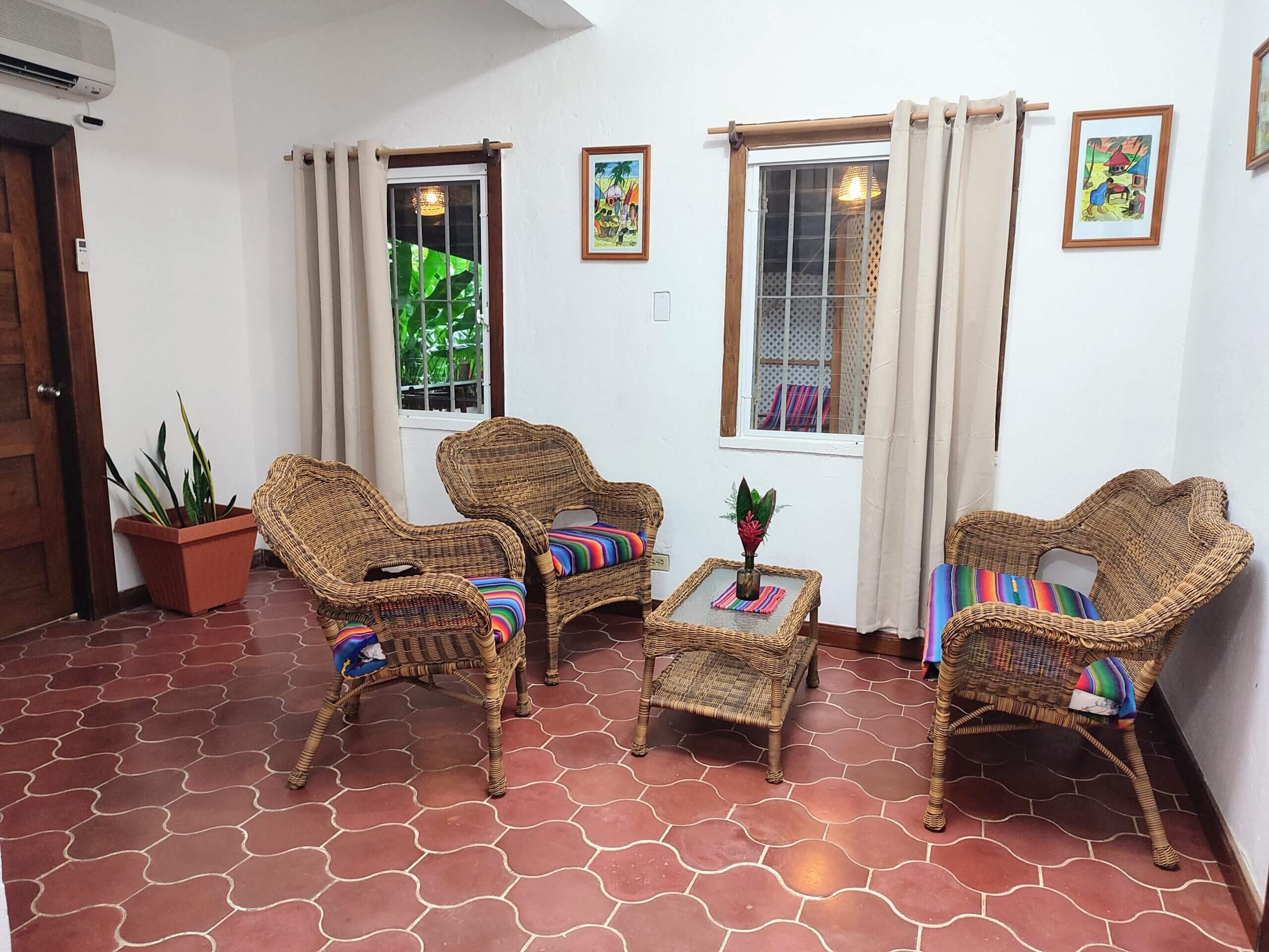 A seating area with red patterned tile flooring, featuring three wicker chairs and a matching coffee table, with neutral tan curtains on the windows and potted snake plants.