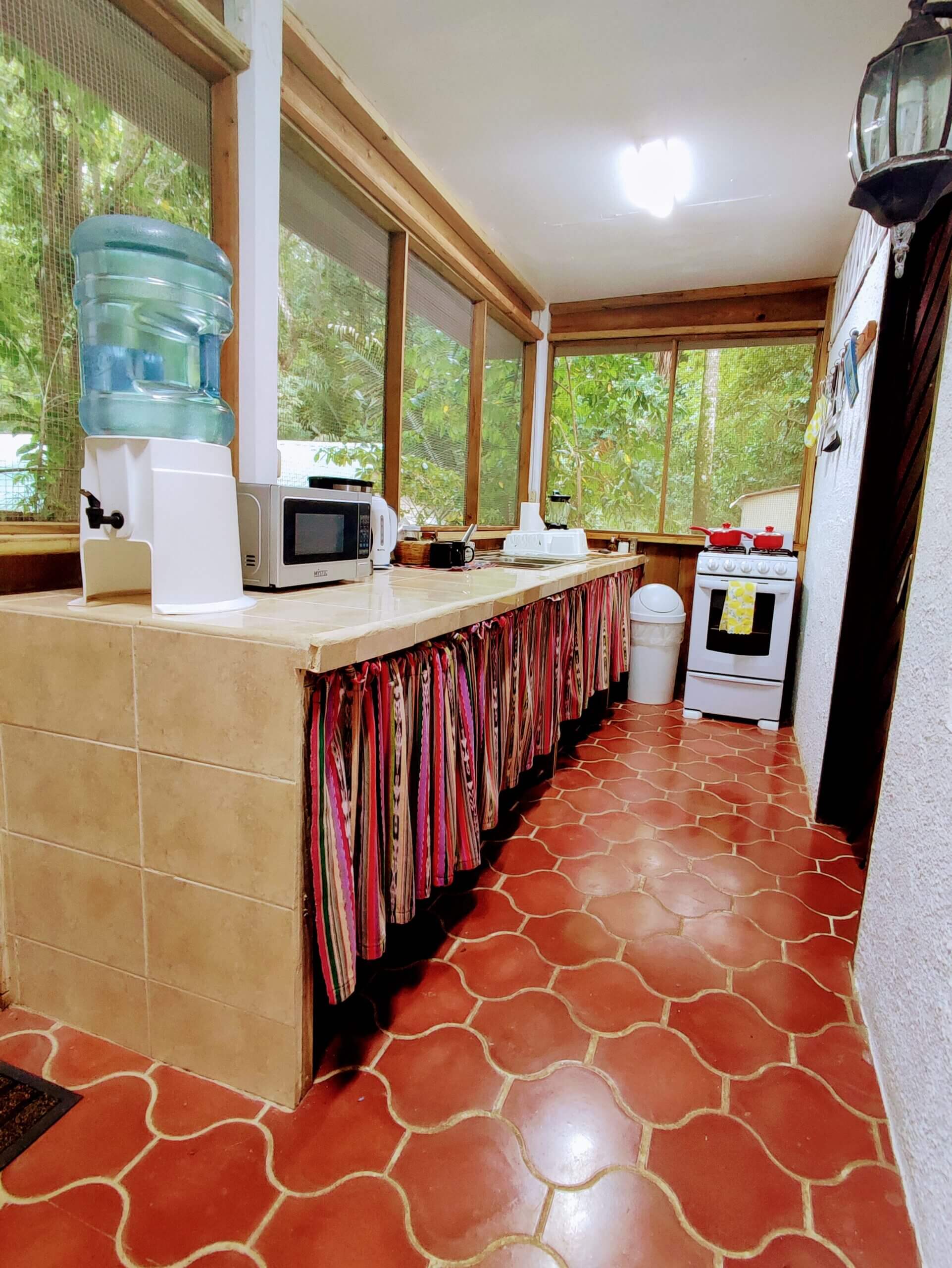 A long, narrow indoor kitchenette with red patterned tile flooring, featuring a long tan counter covered by a striped fabric skirt, a microwave, a water cooler, and a small white stove.