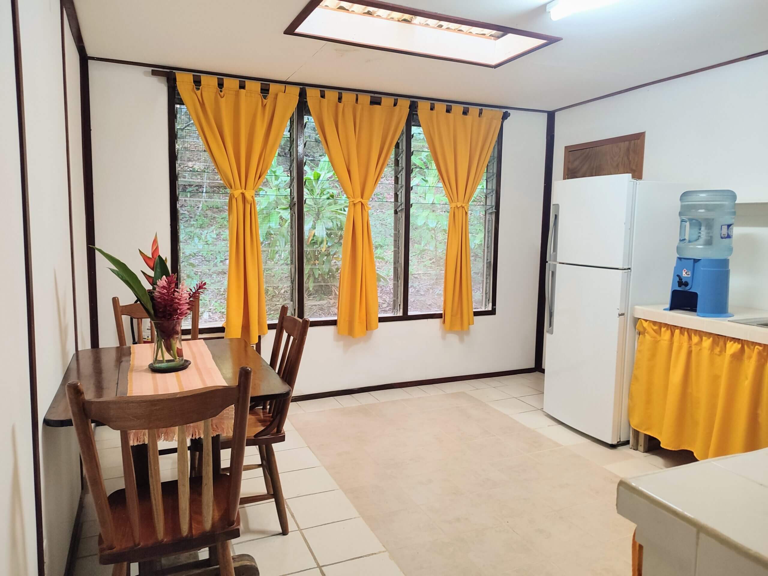A dining and kitchen area with light-colored tile flooring, featuring a wooden table with chairs, a white refrigerator, and windows with bright yellow curtains.