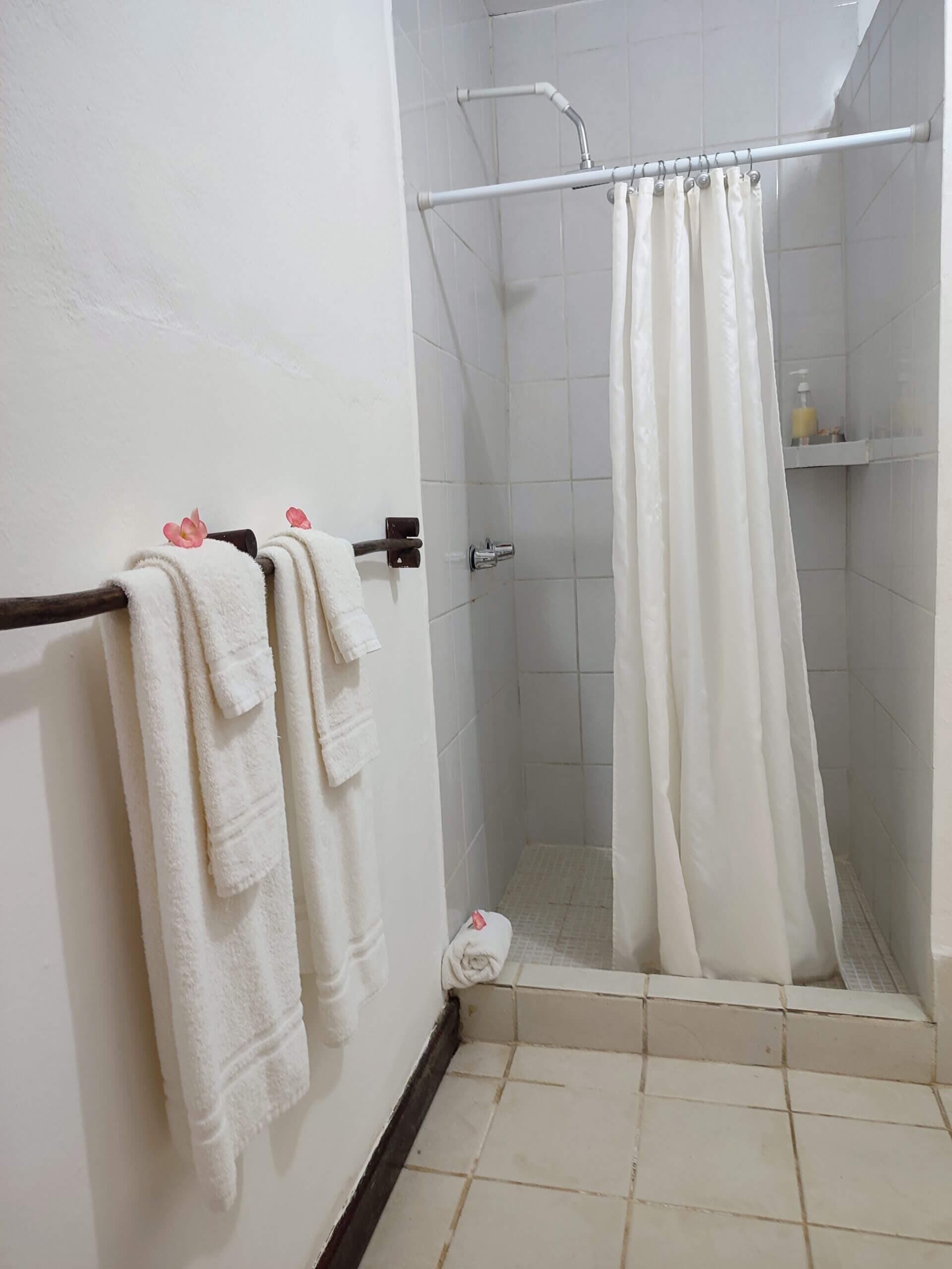 A simple bathroom featuring white walls and light-colored tile flooring, with two white towels hanging on a dark wood rack and a walk-in shower with a white curtain.