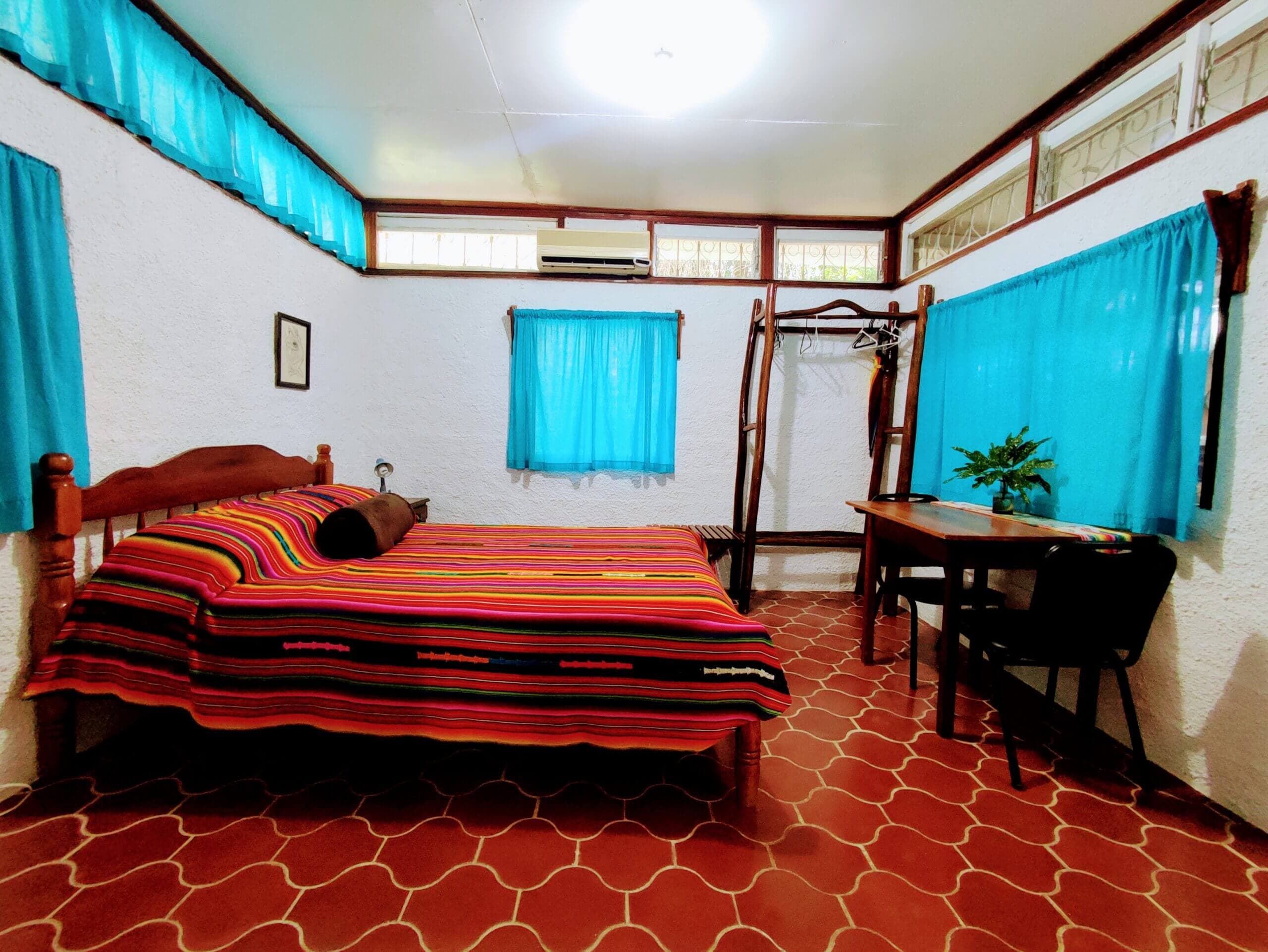 A bedroom with white walls and red patterned tile flooring, featuring a bed with a vibrant multi-colored striped blanket, a wooden dining table with black chairs, and windows with bright teal curtains.