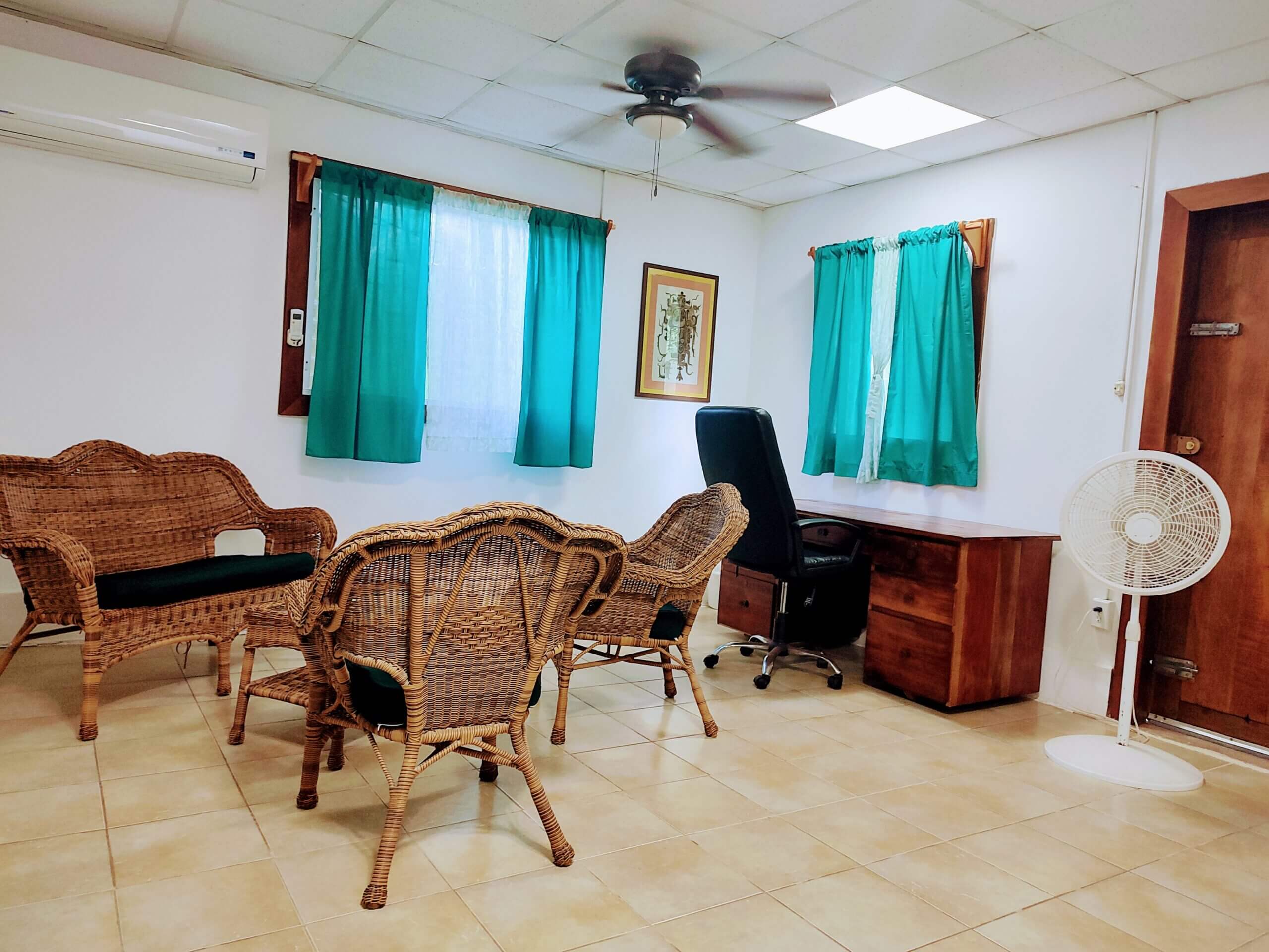 A spacious living or office area with light-colored tile flooring, featuring a set of wicker chairs and a matching sofa, a large wooden desk with a black office chair, a white pedestal fan, and windows with bright teal curtains.