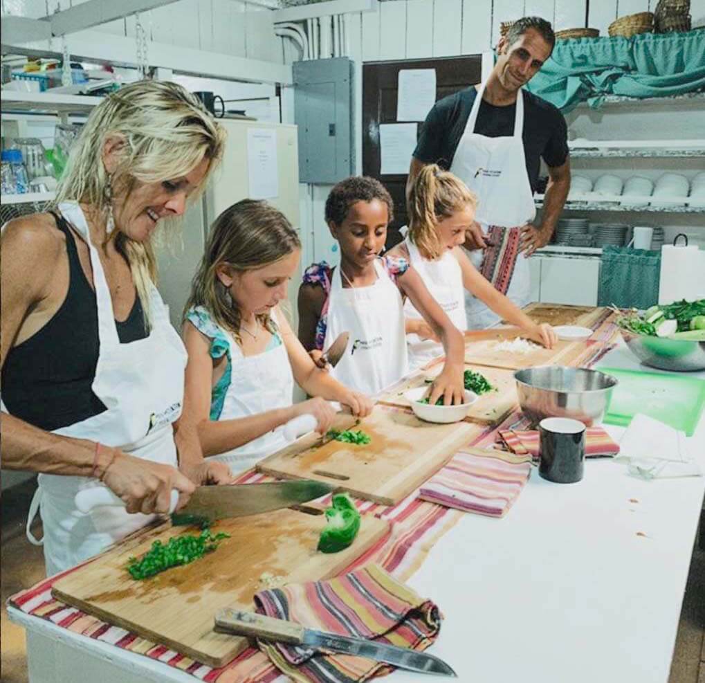 A group of four individuals, including children and an adult, are preparing food in an indoor kitchen, chopping vegetables on wooden boards.