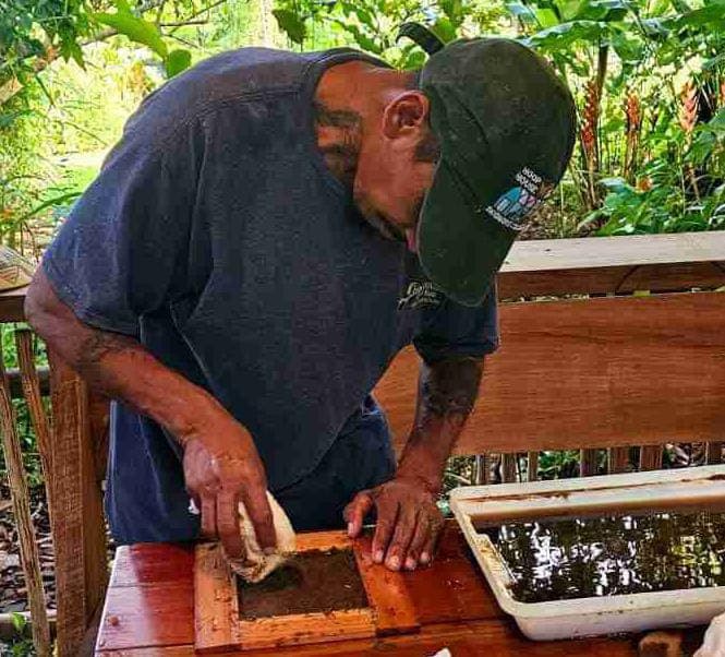 A man in a cap works on a wooden frame filled with dark material at a table surrounded by greenery.