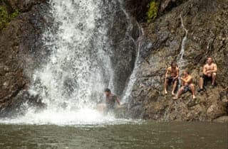 A group of four people sits by a waterfall, with one person jumping into the water below.