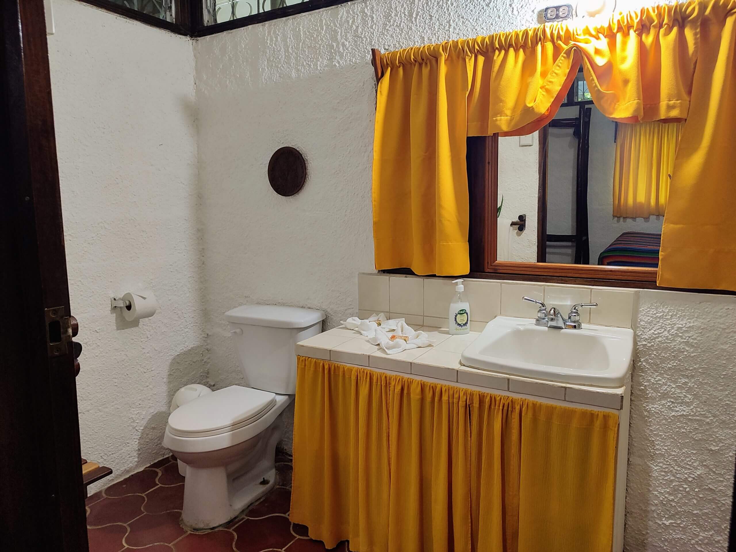 A bathroom featuring a white vanity with a bright yellow fabric skirt and matching yellow curtains over a mirror, a white toilet, and red patterned tile flooring.