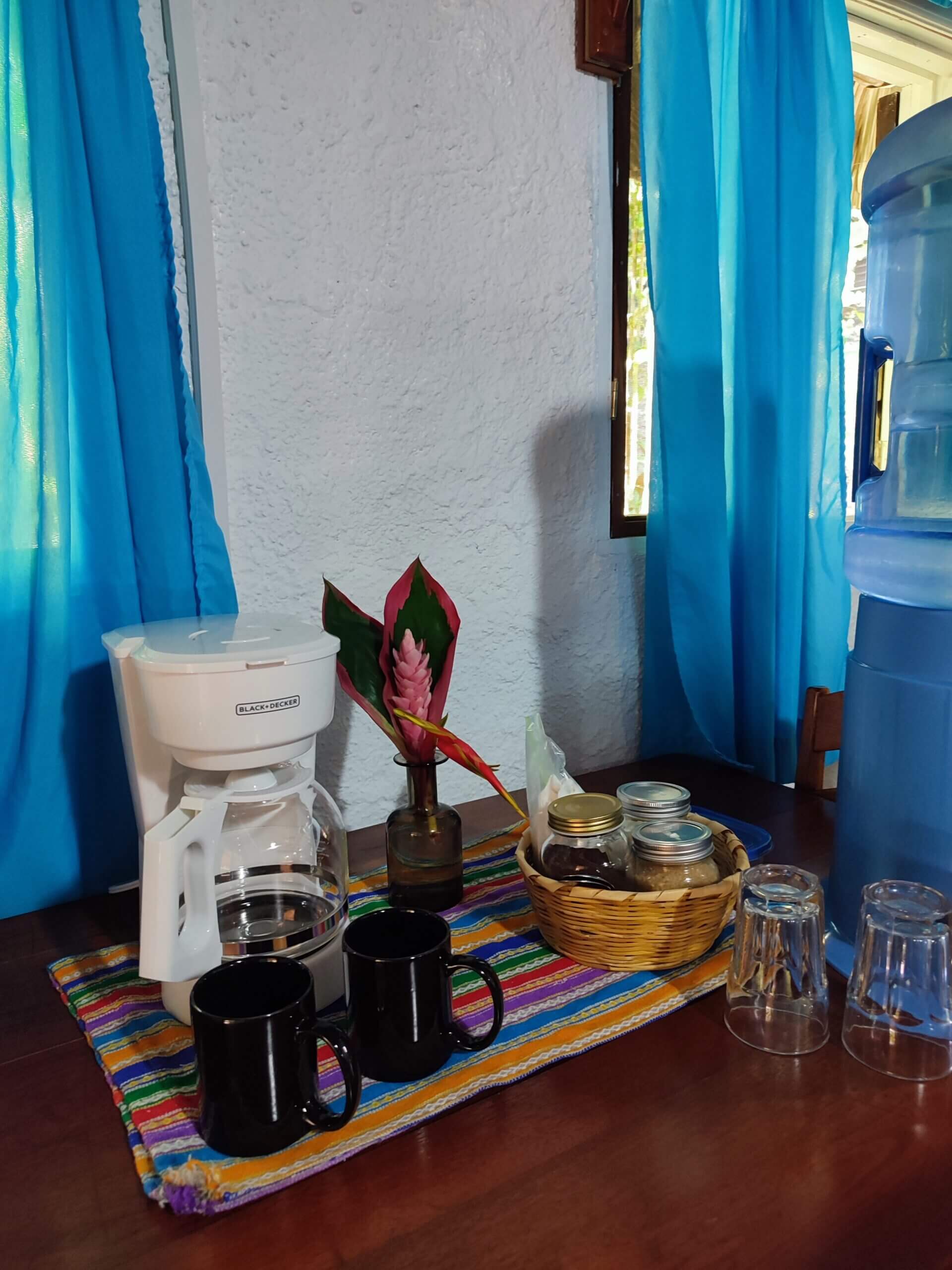 A close-up shot of a wooden surface featuring a white coffee maker, two black mugs, a small basket of supplies, and clear glasses set on a vibrant multi-colored striped mat, positioned between windows with bright blue curtains.