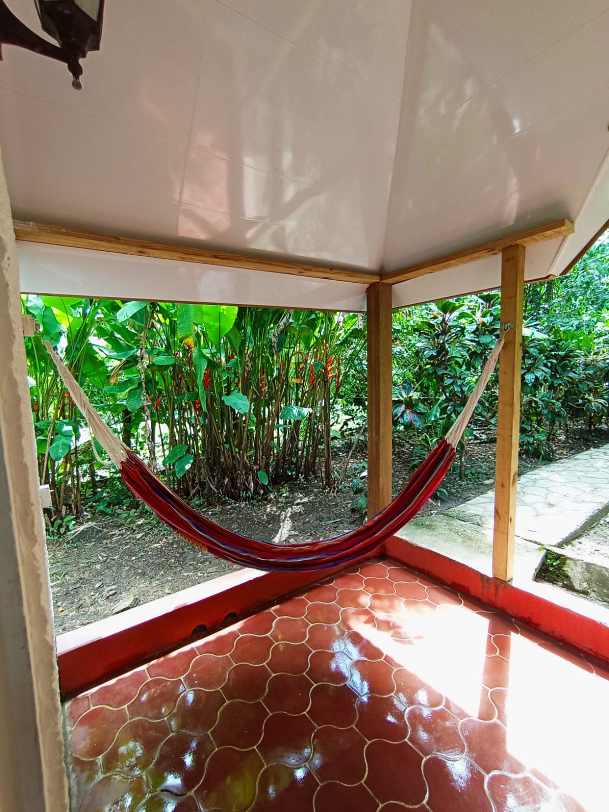 A red hammock hangs on a covered porch with red patterned tile flooring, looking out onto a lush garden of tropical plants and green foliage.