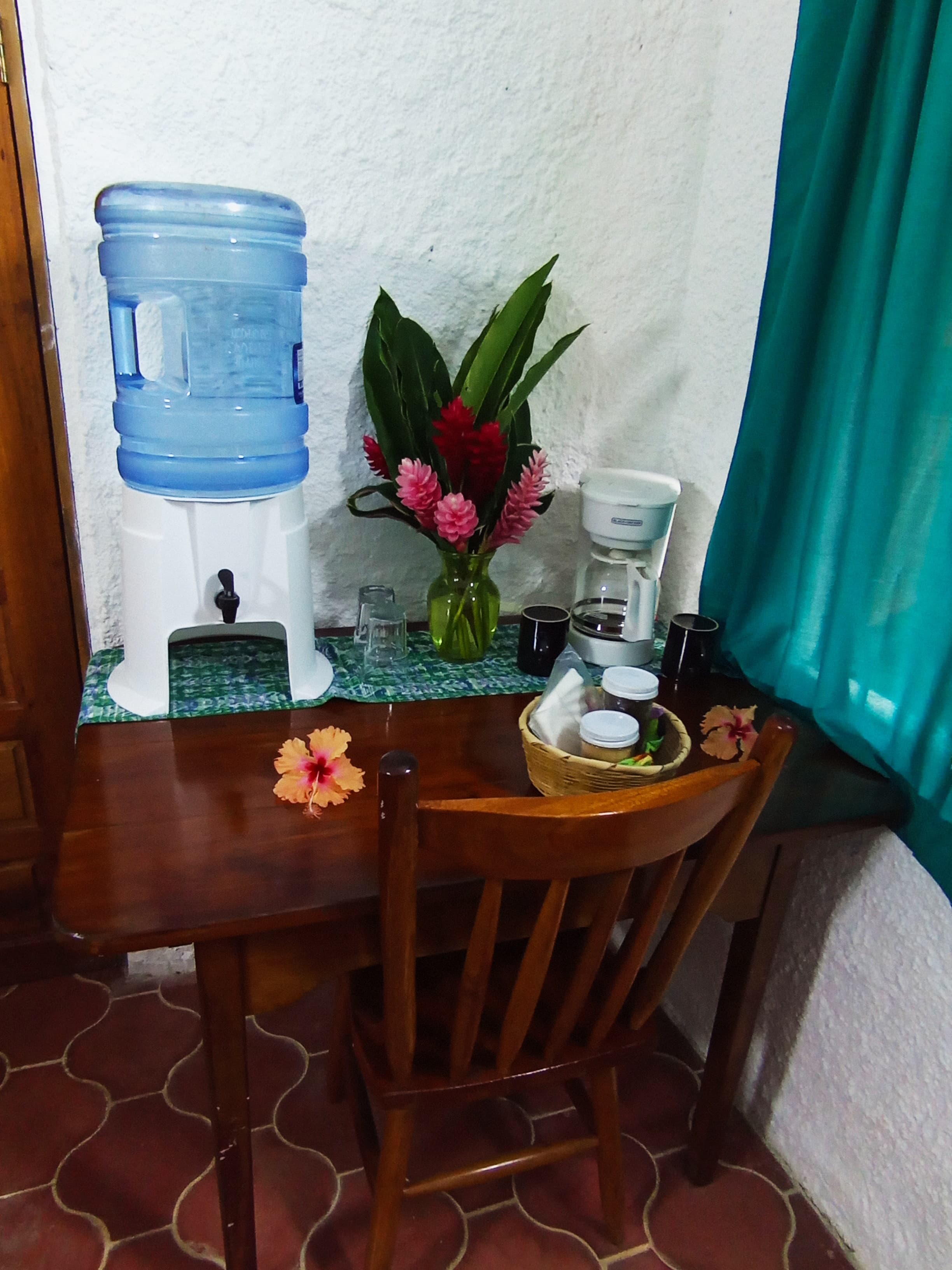 An indoor wooden table set with a water cooler, coffee maker, and fresh tropical flowers, accompanied by a single wooden chair against a white wall.