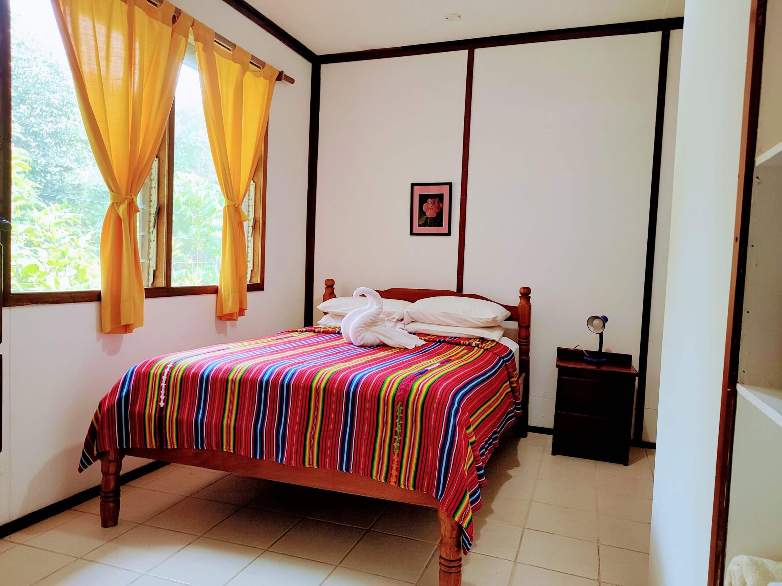 A bedroom with white paneled walls and a wooden bed featuring a vibrant, multi-colored striped blanket, two pillows, and a window with bright yellow curtains.