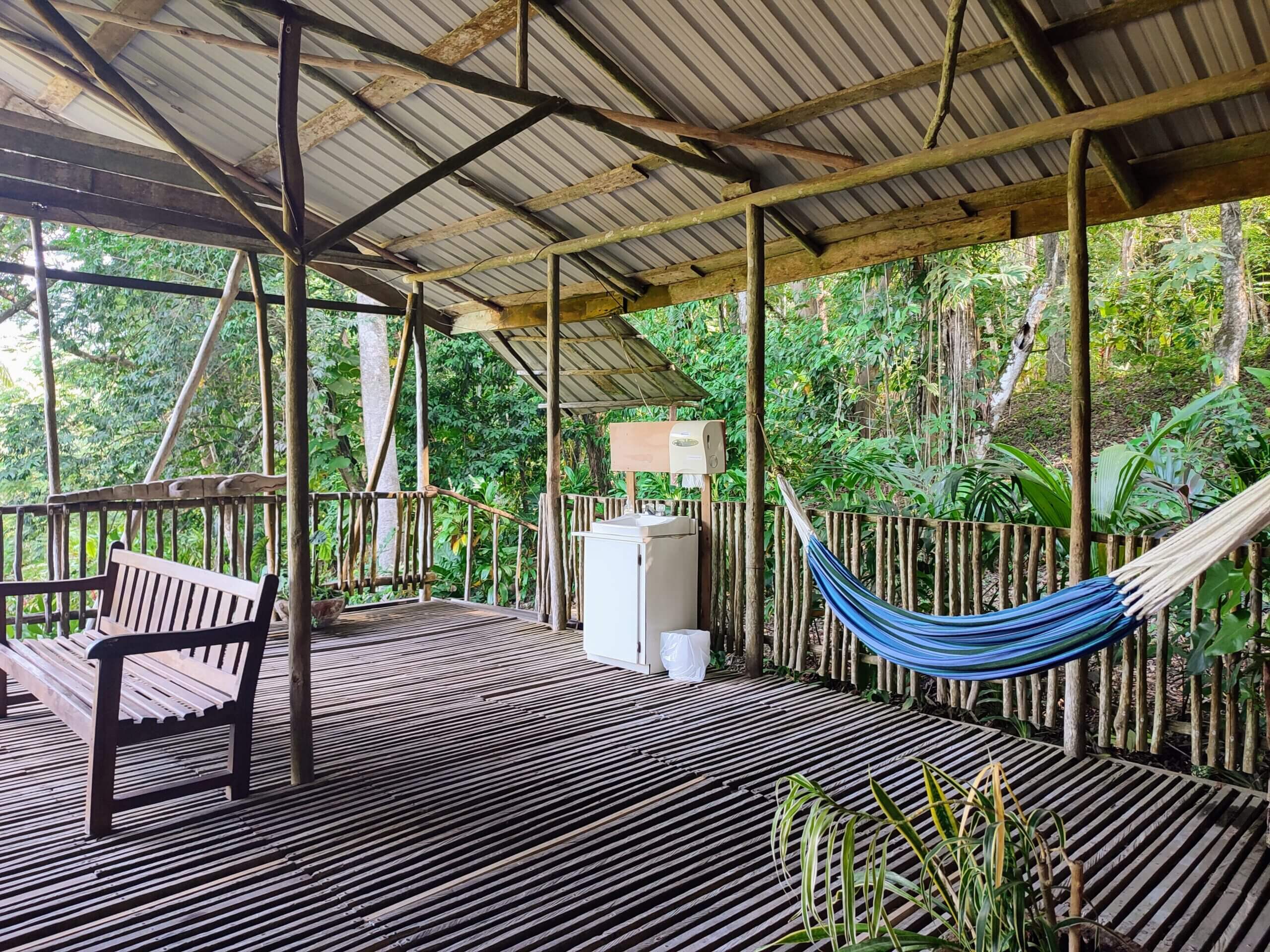 A spacious wooden deck under a corrugated metal roof, featuring a wooden bench, a blue hammock, surrounded by a rustic wooden railing and lush green forest.