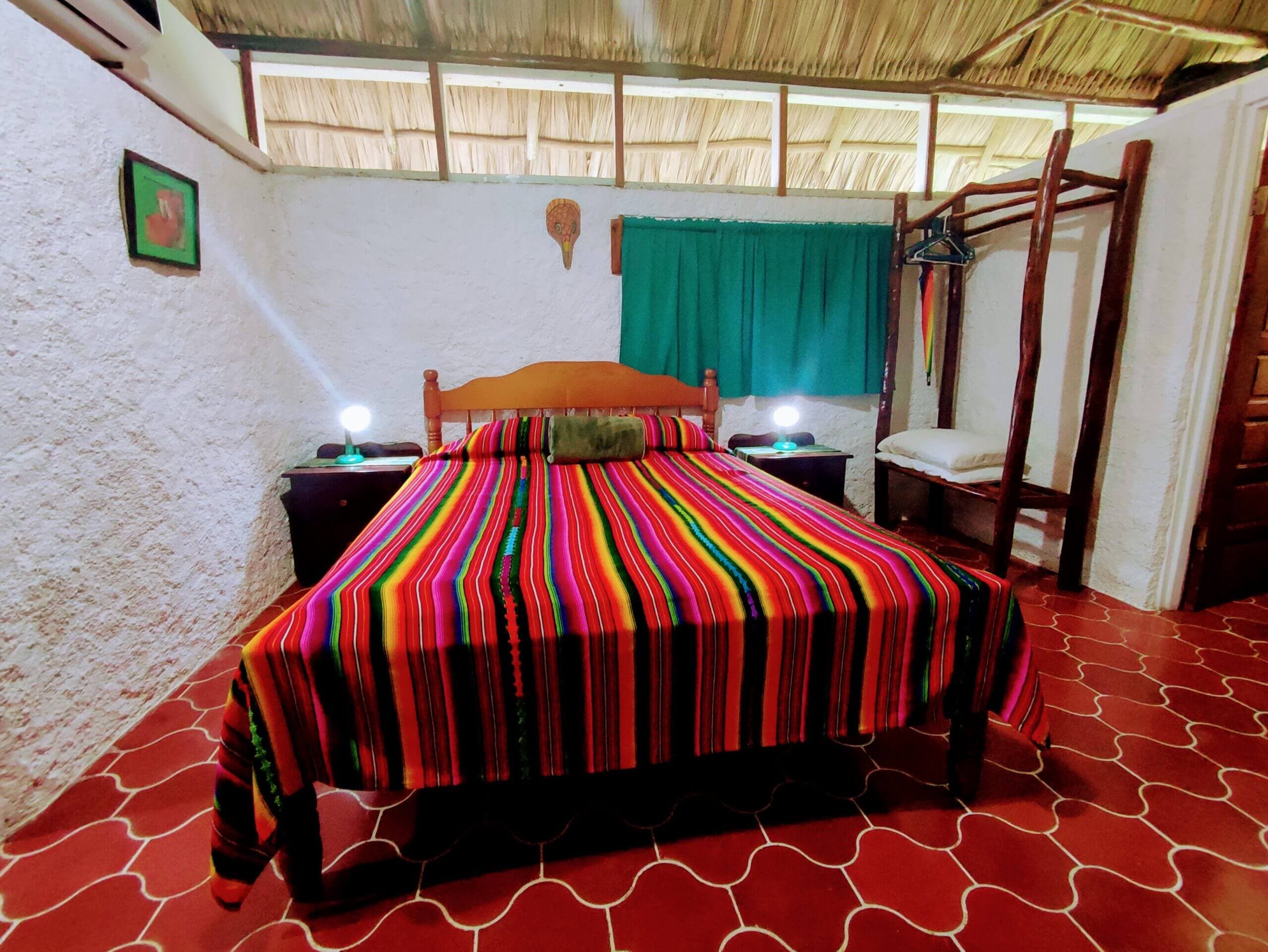 A bedroom with red patterned tile flooring and a vaulted wooden ceiling, featuring a large bed with a vibrant multi-colored striped blanket, dark wood nightstands, and a window with deep green curtains.