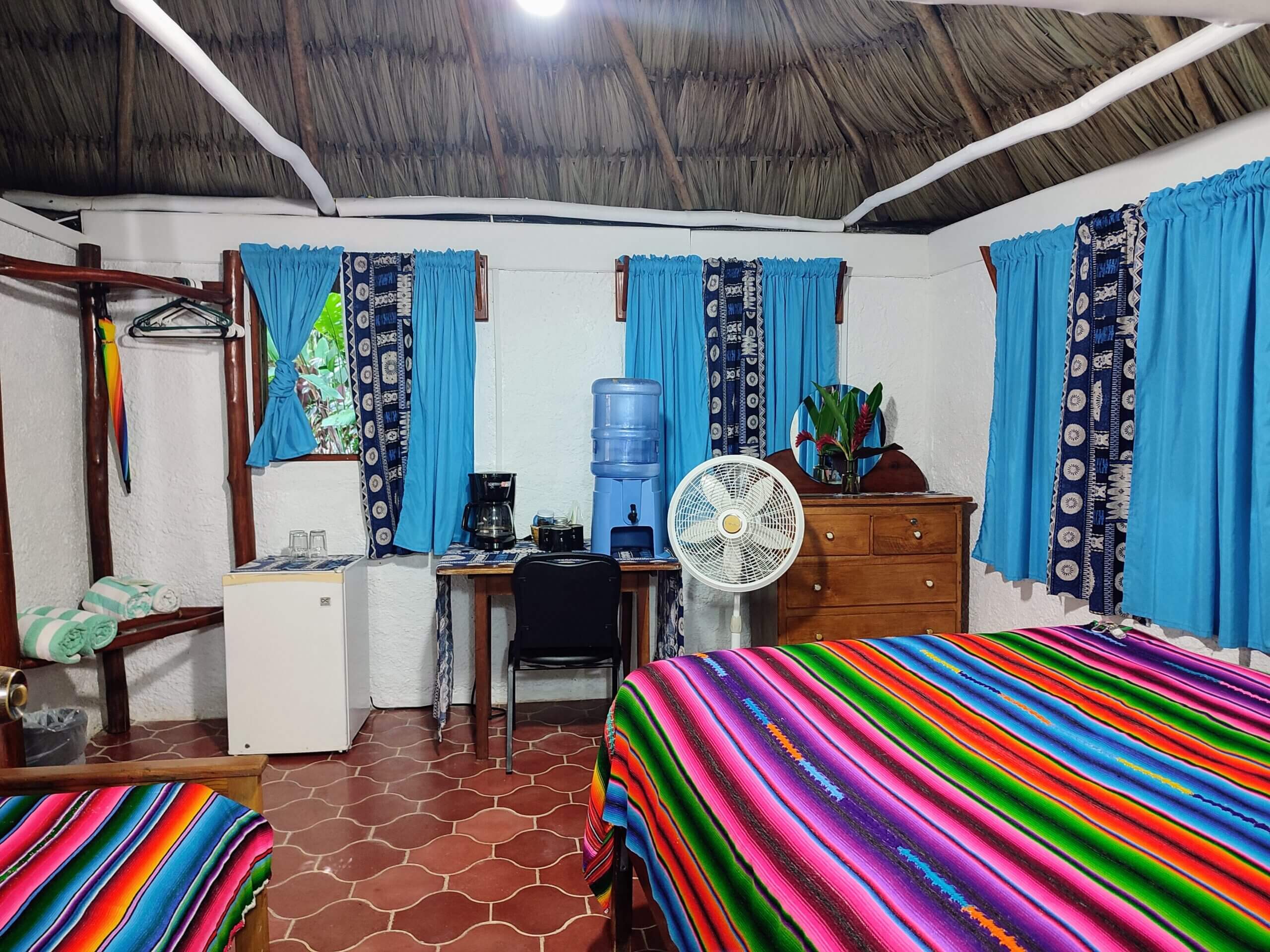 A bedroom with white walls and red patterned tile flooring, featuring a bed with a vibrant multi-colored striped blanket, a small refrigerator, a desk with a chair, and windows with bright blue curtains.