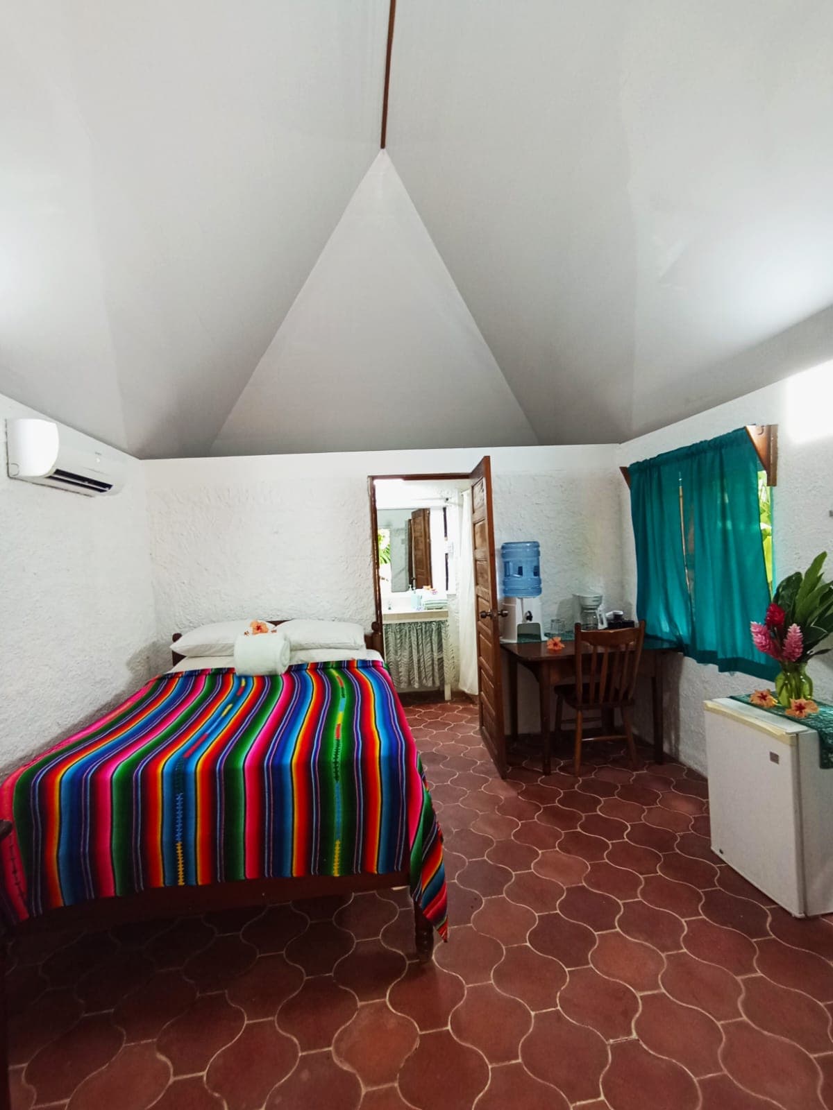 A bedroom with white walls, red patterned tile flooring, and a vaulted ceiling, featuring a bed with a vibrant multi-colored striped blanket and a small wooden desk under a teal curtained window.