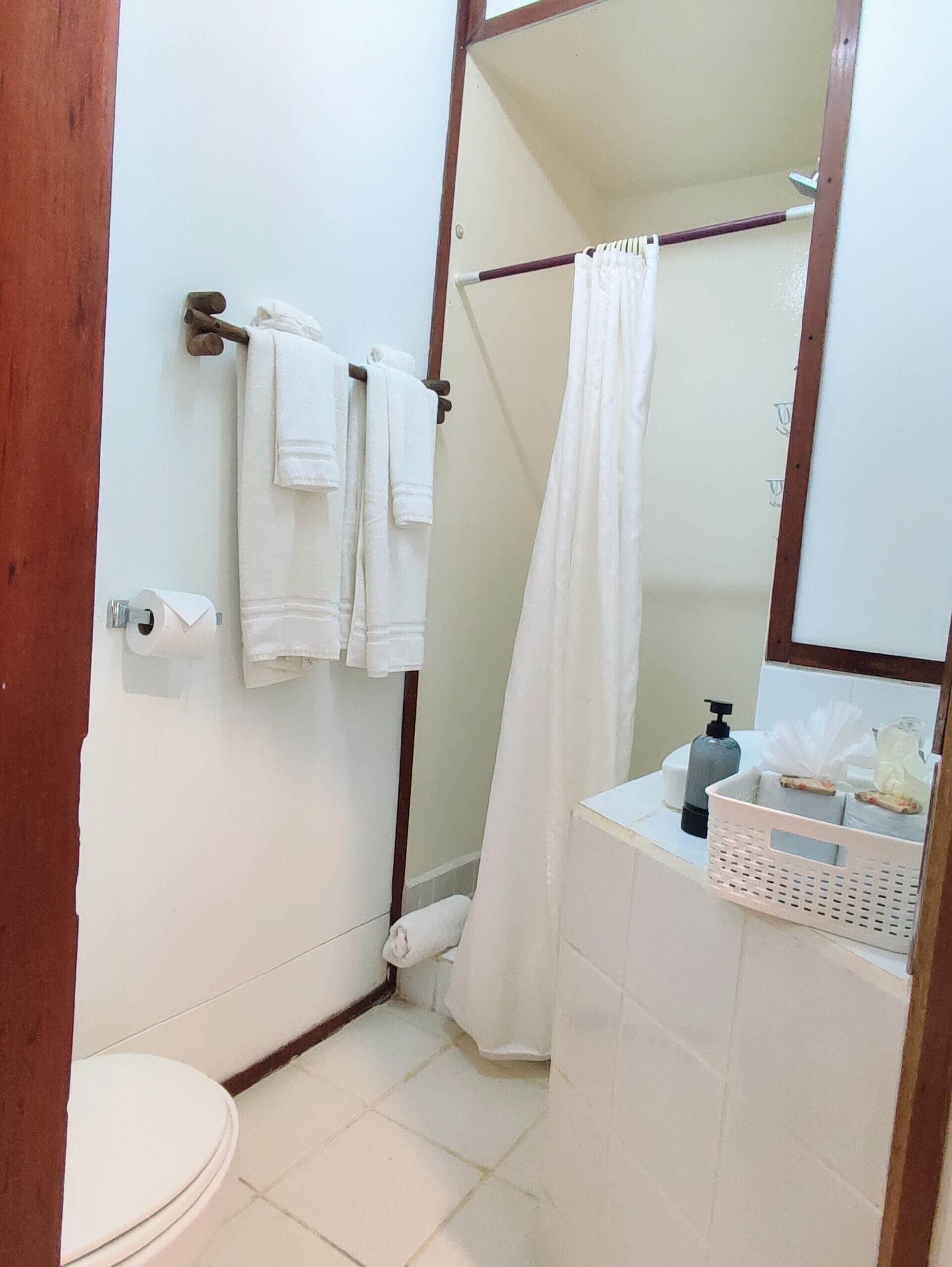 A bathroom featuring a white vanity, a toilet, and white towels hanging on a dark rack, with a white shower curtain visible in the background against light-colored tile flooring.