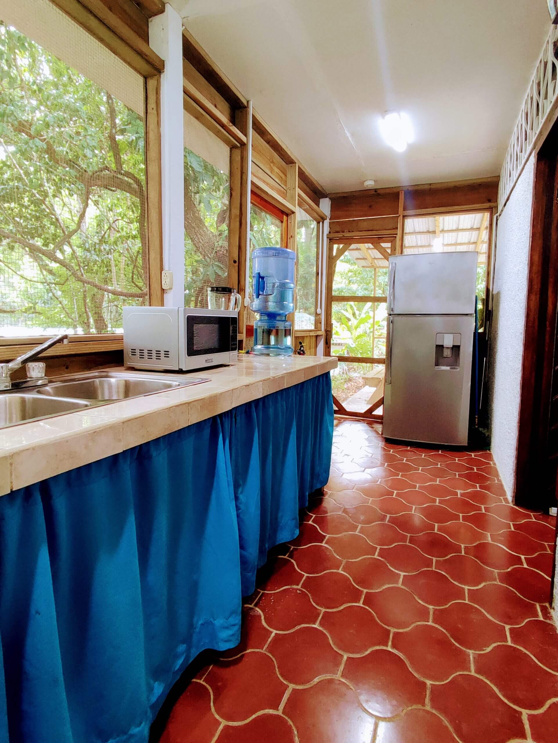 A long, narrow indoor kitchenette with red patterned tile flooring, featuring a stainless steel sink, a microwave, and a water cooler on a tan counter with a bright blue fabric skirt, leading toward a refrigerator at the end of the hall.