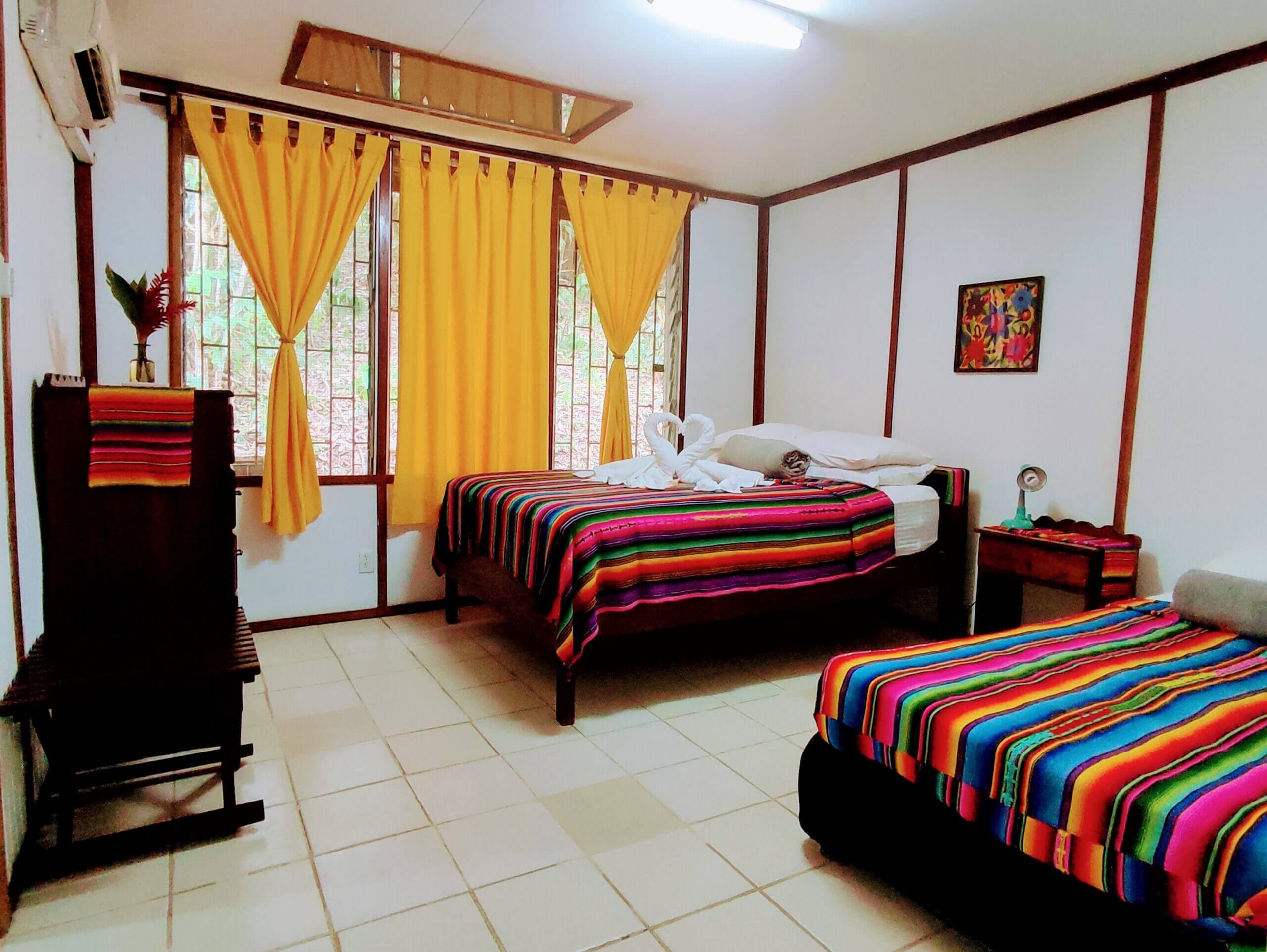 A bedroom with white paneled walls and light-colored tile flooring, featuring two beds covered in vibrant multi-colored striped blankets and windows with bright yellow curtains.