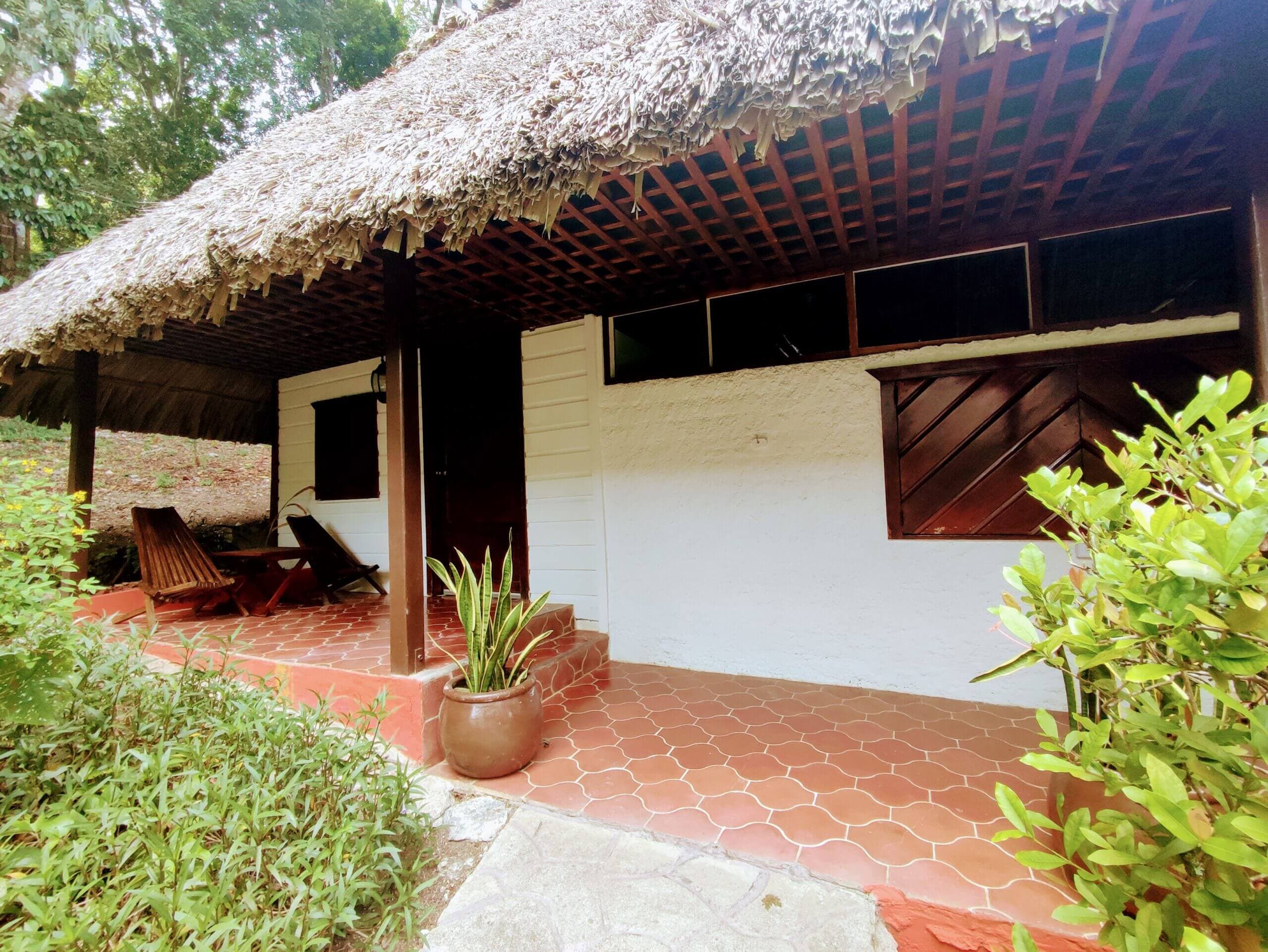 An exterior view of a white building with a large thatched roof, featuring a shaded porch with red patterned tile flooring, wooden lounge chairs, and a potted plant.